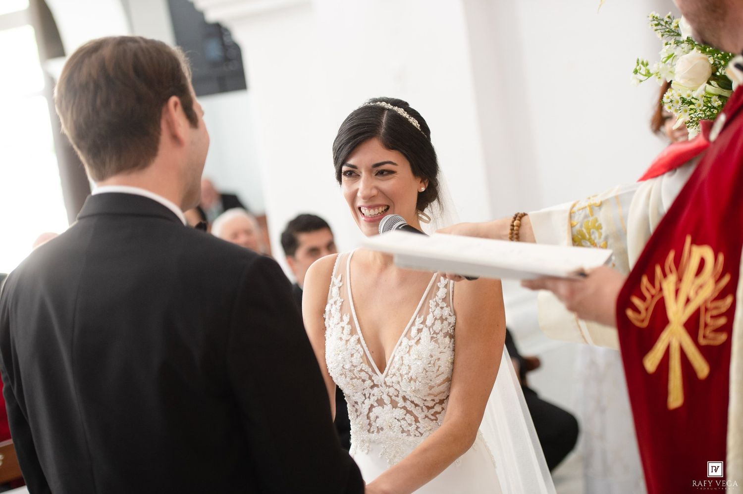 Bride smiling joyfully during wedding ceremony in church, wearing lace gown and bridal headpiece, facing groom.