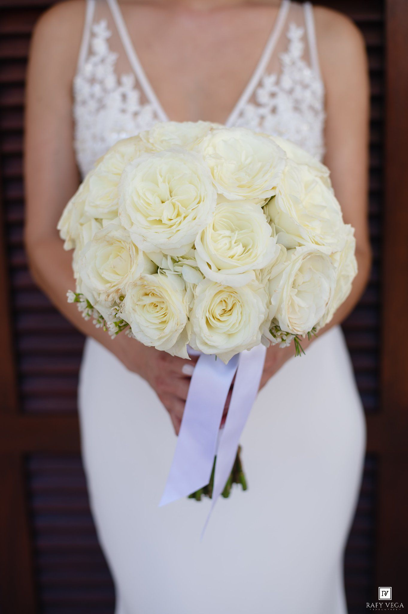 Bride in white lace gown holding elegant round bouquet of cream roses tied with white ribbon.