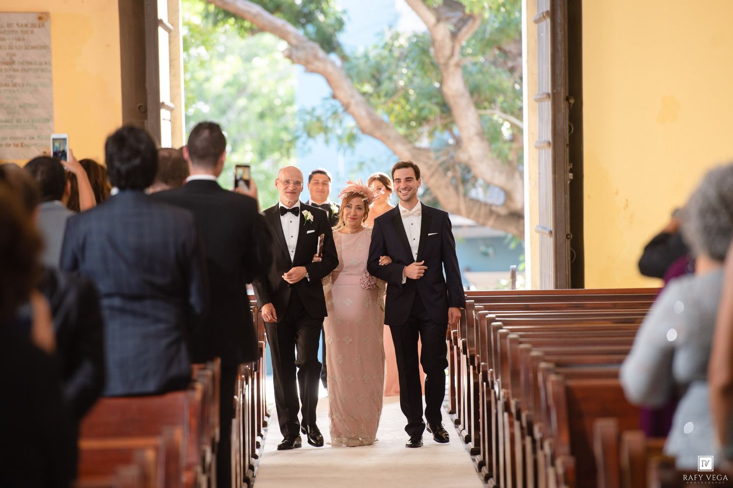 Bride walks down church aisle escorted by two men in suits during a elegant wedding ceremony.