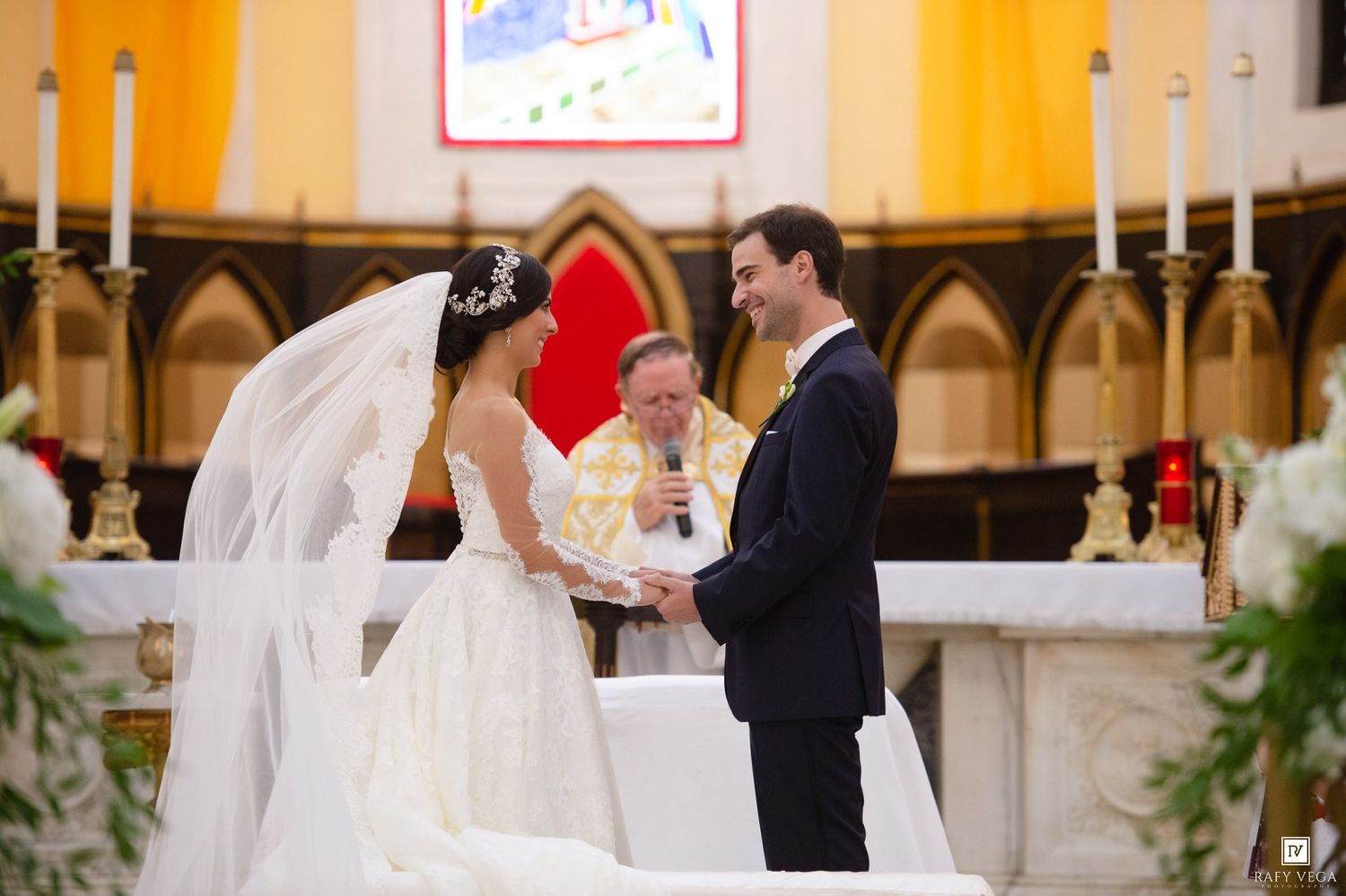 Bride and groom hold hands during wedding ceremony as officiant speaks at ornate church altar with candles.