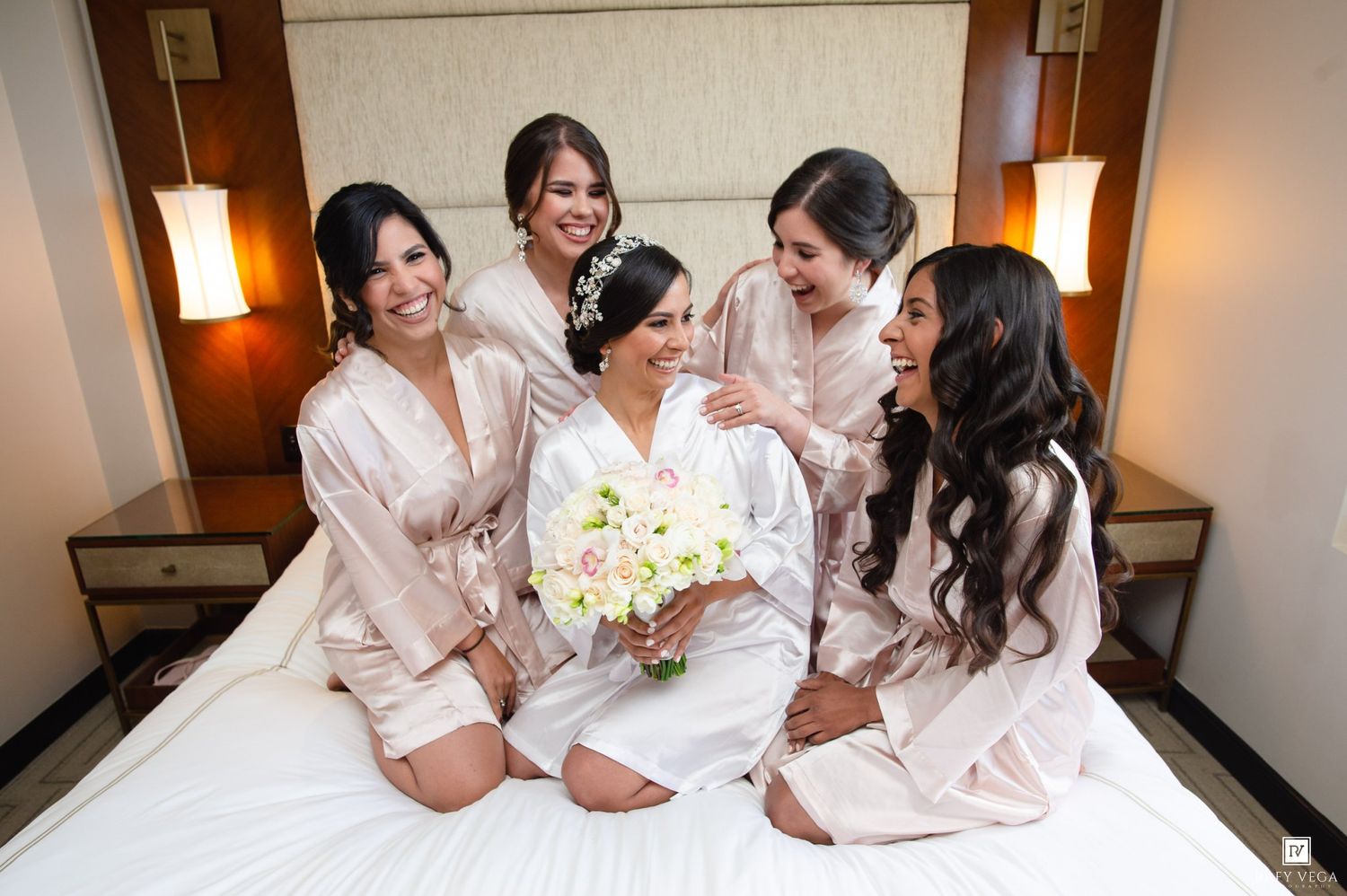 Bride in white robe holding bouquet sits on bed surrounded by smiling bridesmaids in blush robes in hotel room.