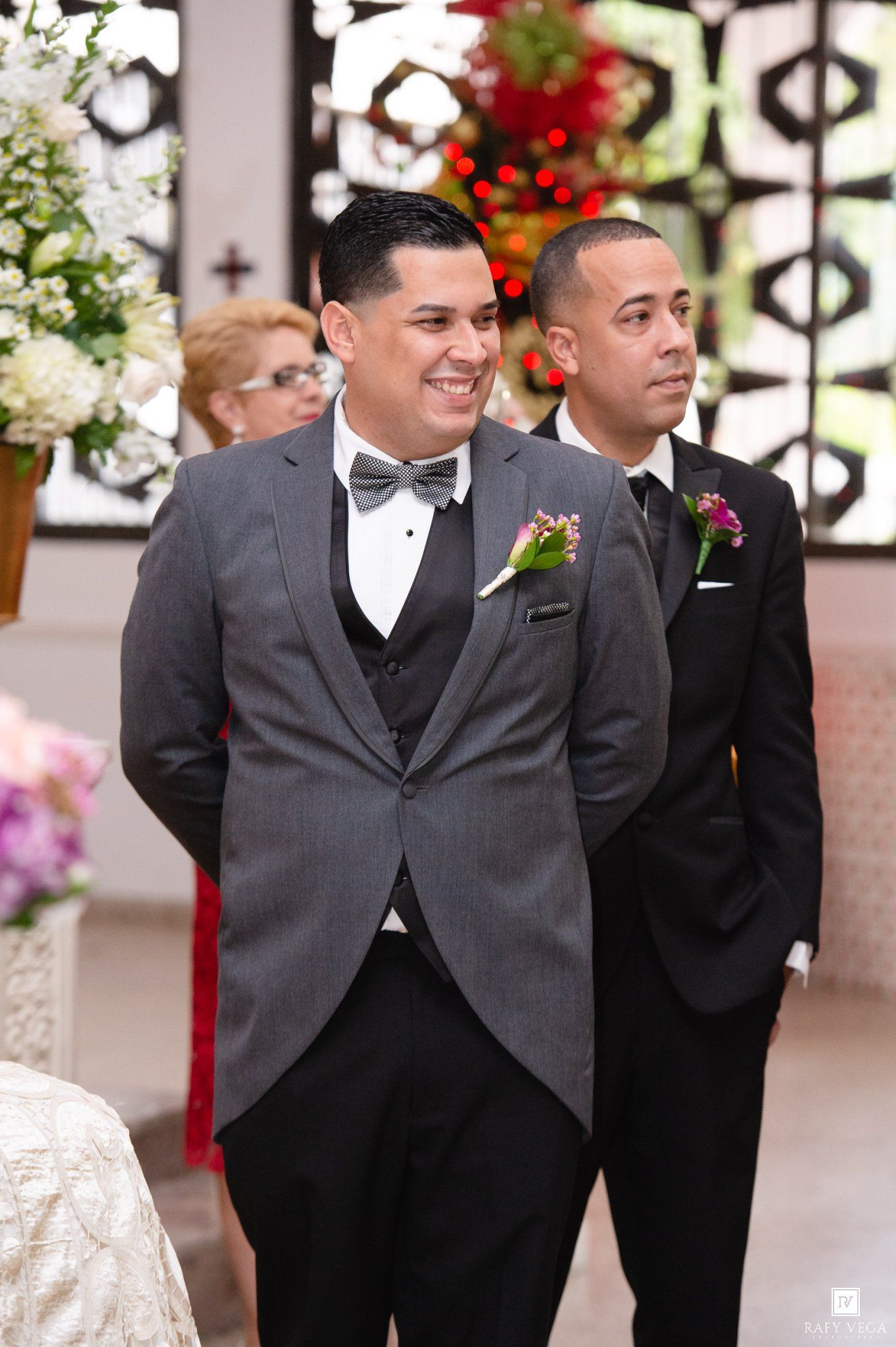 Smiling groom in gray suit with boutonniere stands at altar during wedding ceremony.