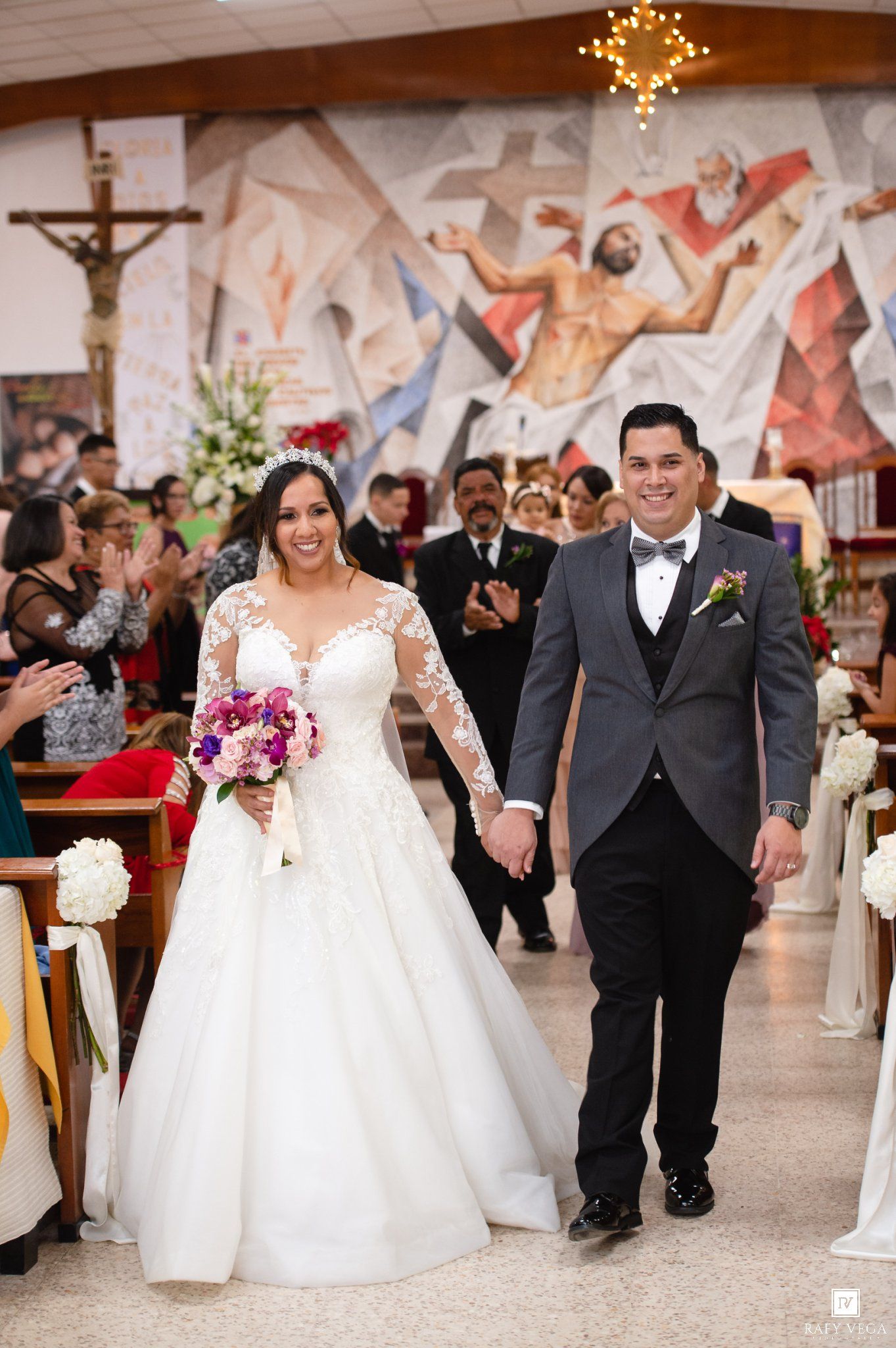 Bride in lace gown and groom in gray tuxedo walk down church aisle after wedding ceremony.