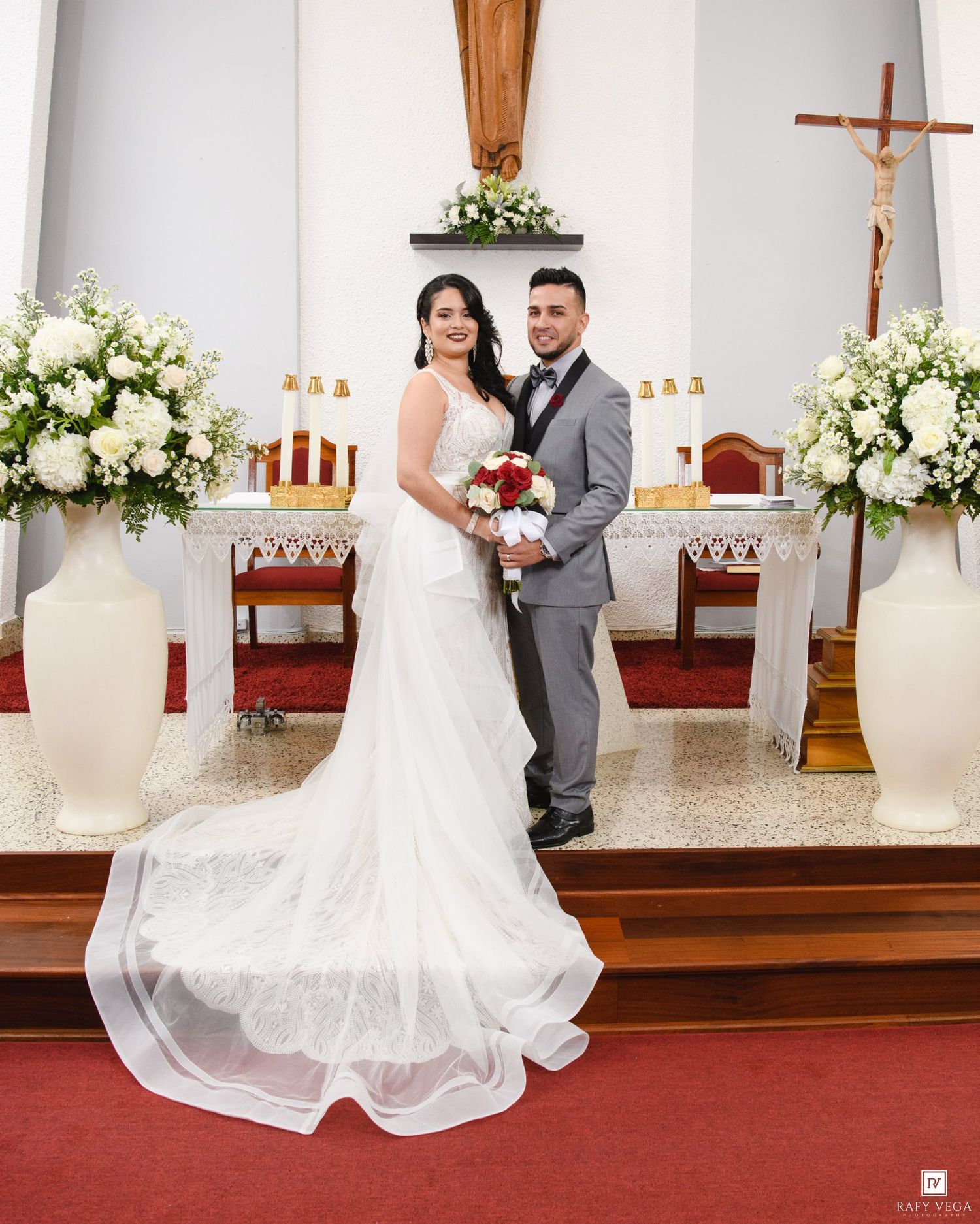 Bride in white gown and groom in gray suit pose at church altar with white floral arrangements and crucifix.