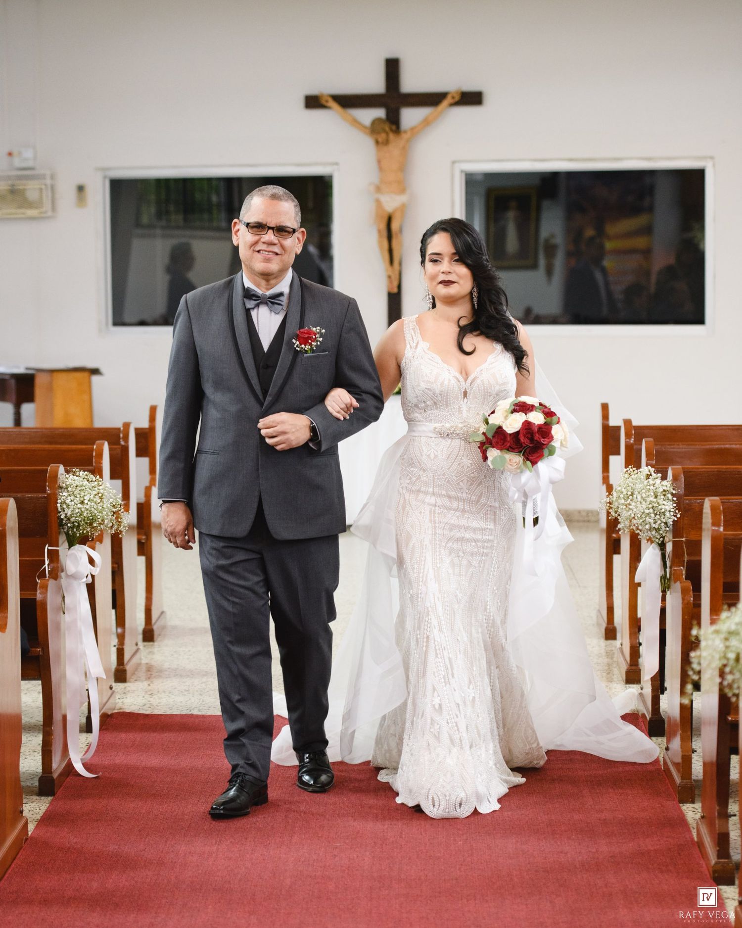 Bride in white lace gown holding red bouquet walks down church aisle with escort in gray suit.