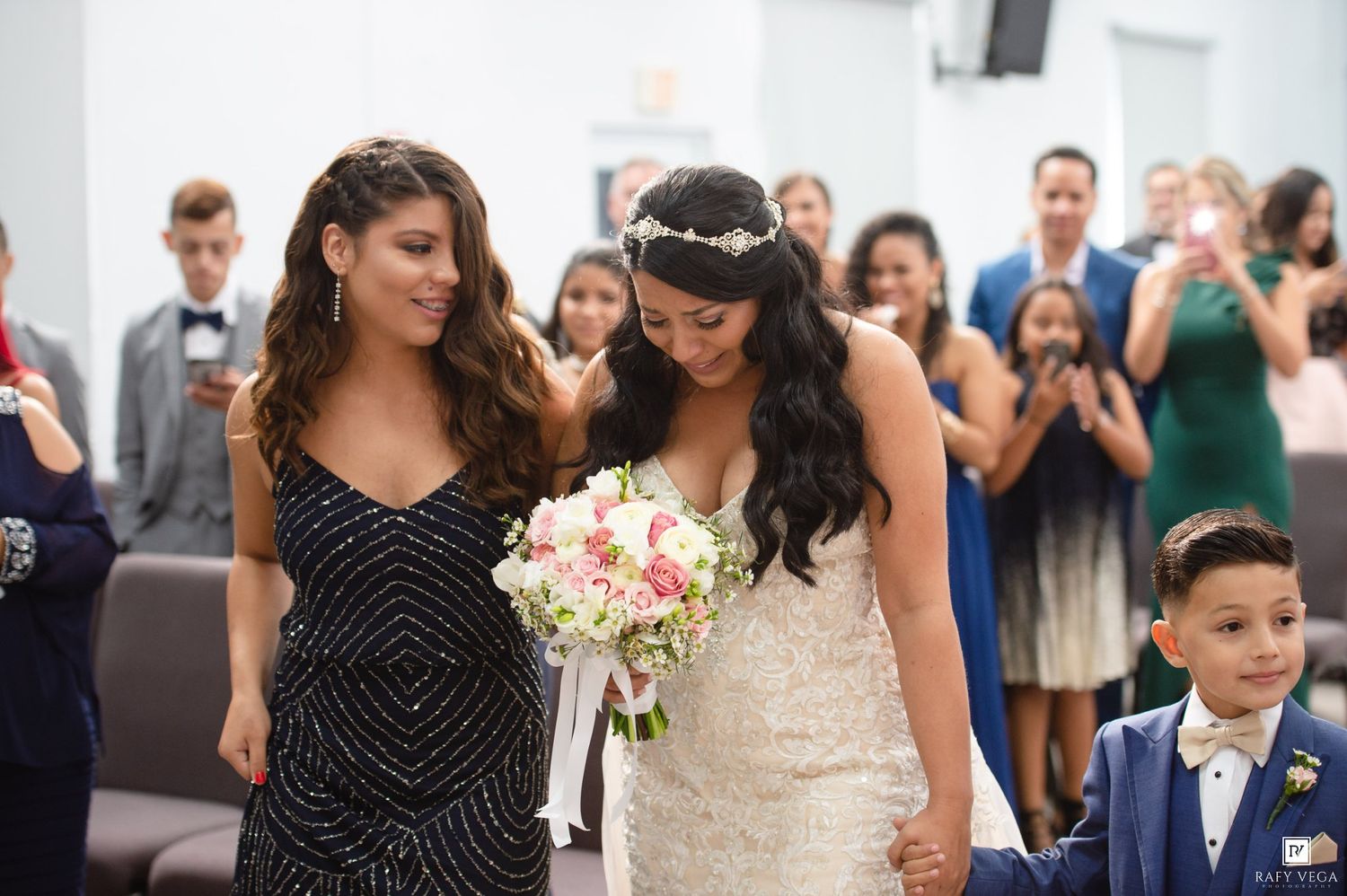 Bride in white gown walks down aisle with bridesmaid in black dress, holding pink bouquet, as guests celebrate.