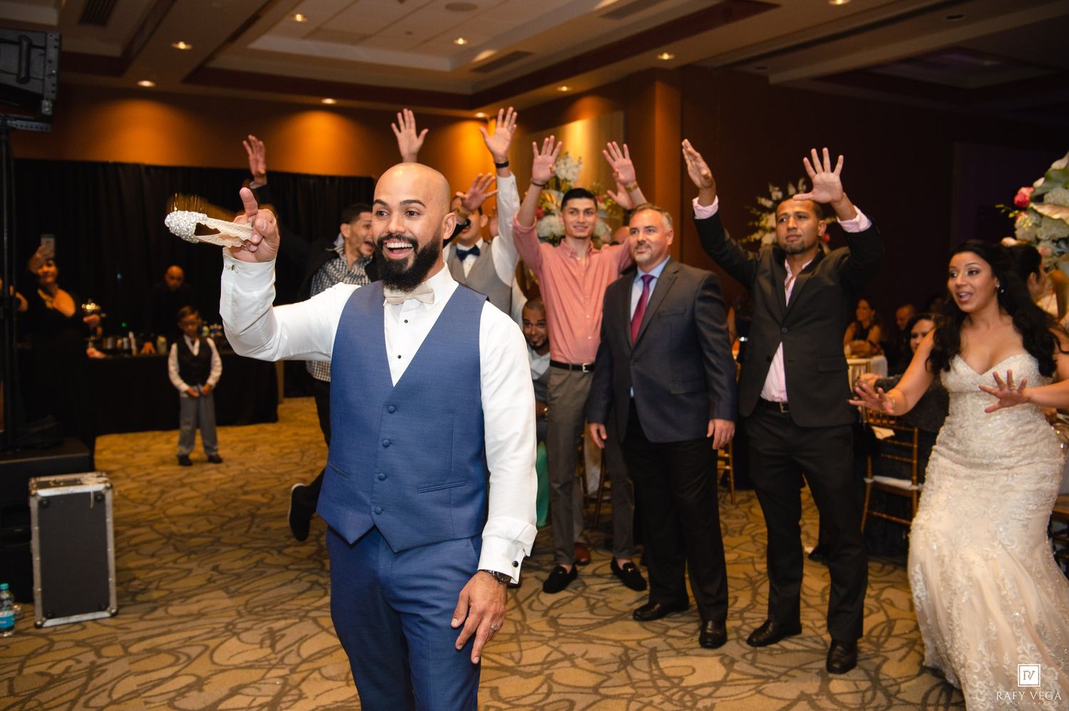 Groom in blue vest celebrates with wedding guests raising hands in a decorated banquet hall reception.