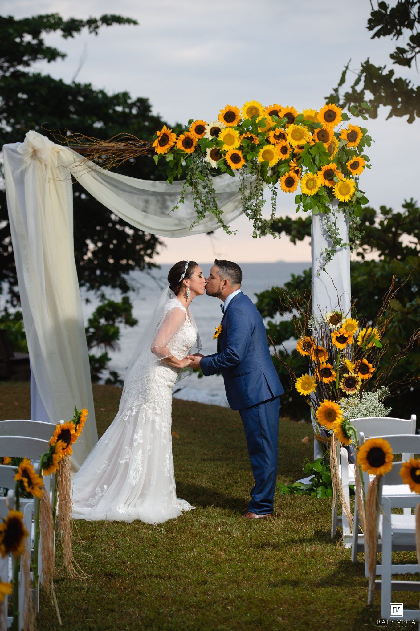 Bride and groom share first kiss under sunflower arch at outdoor waterfront wedding ceremony.