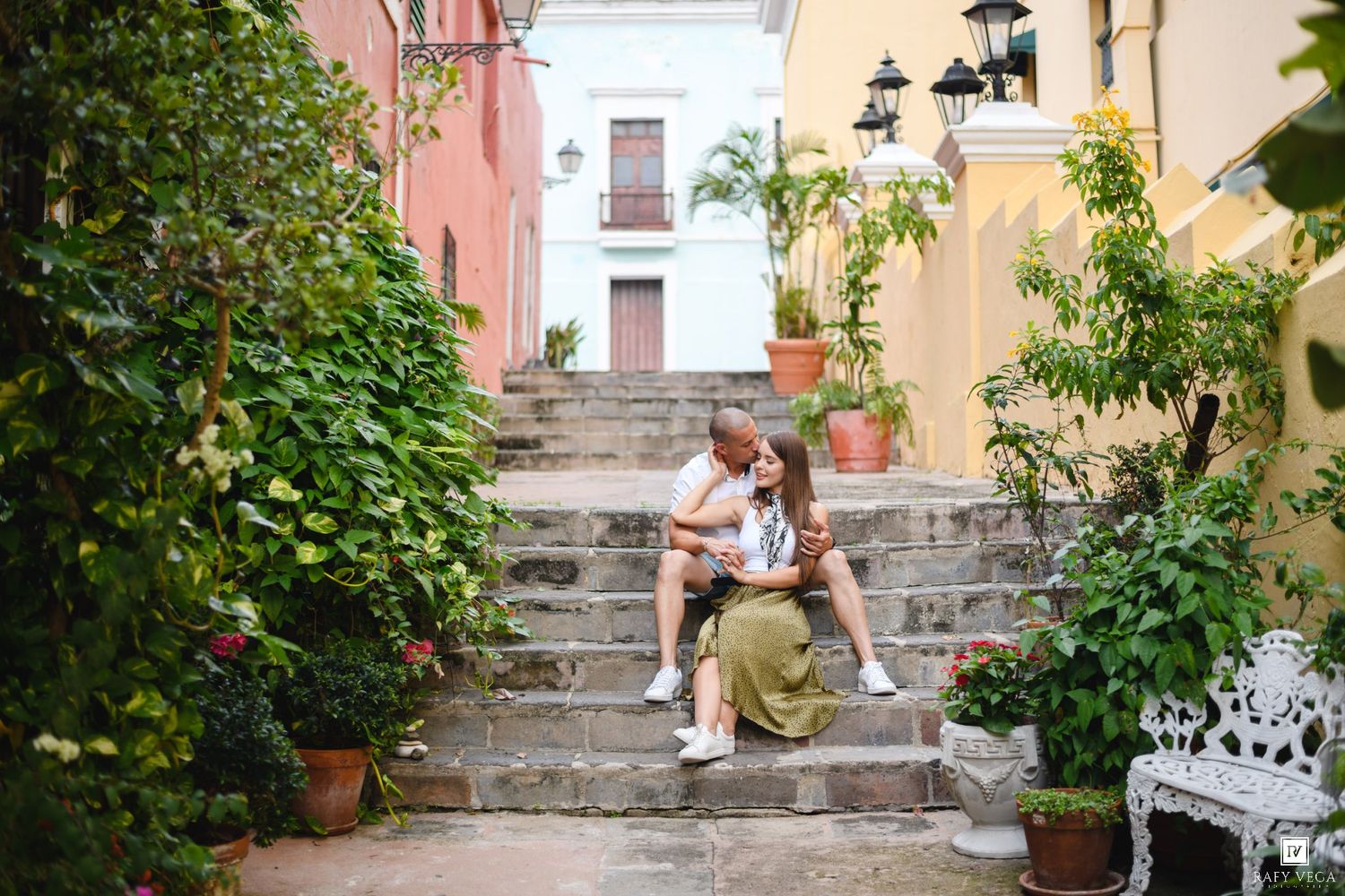 Couple sitting on stone steps surrounded by lush greenery and colorful colonial buildings in a charming outdoor setting.