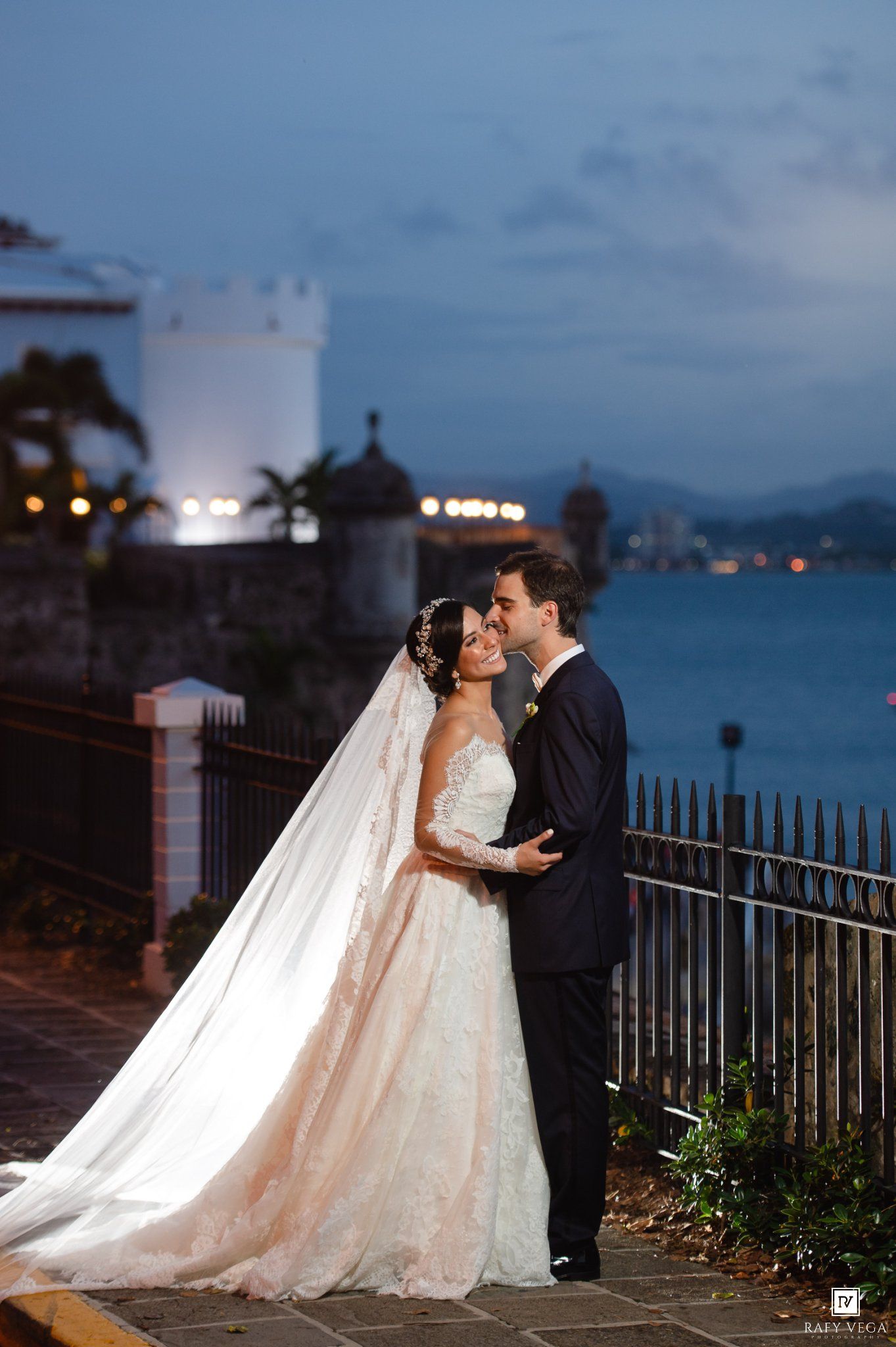 Bride and groom share a romantic kiss at dusk by a waterfront, bride in lace gown with long veil.