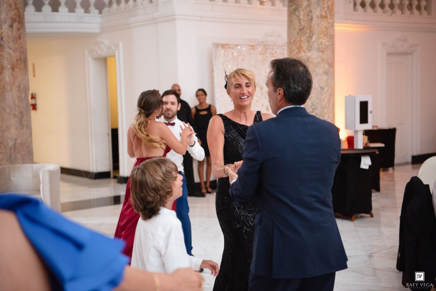 Elegantly dressed guests socializing and dancing at a formal wedding reception in a grand marble ballroom.