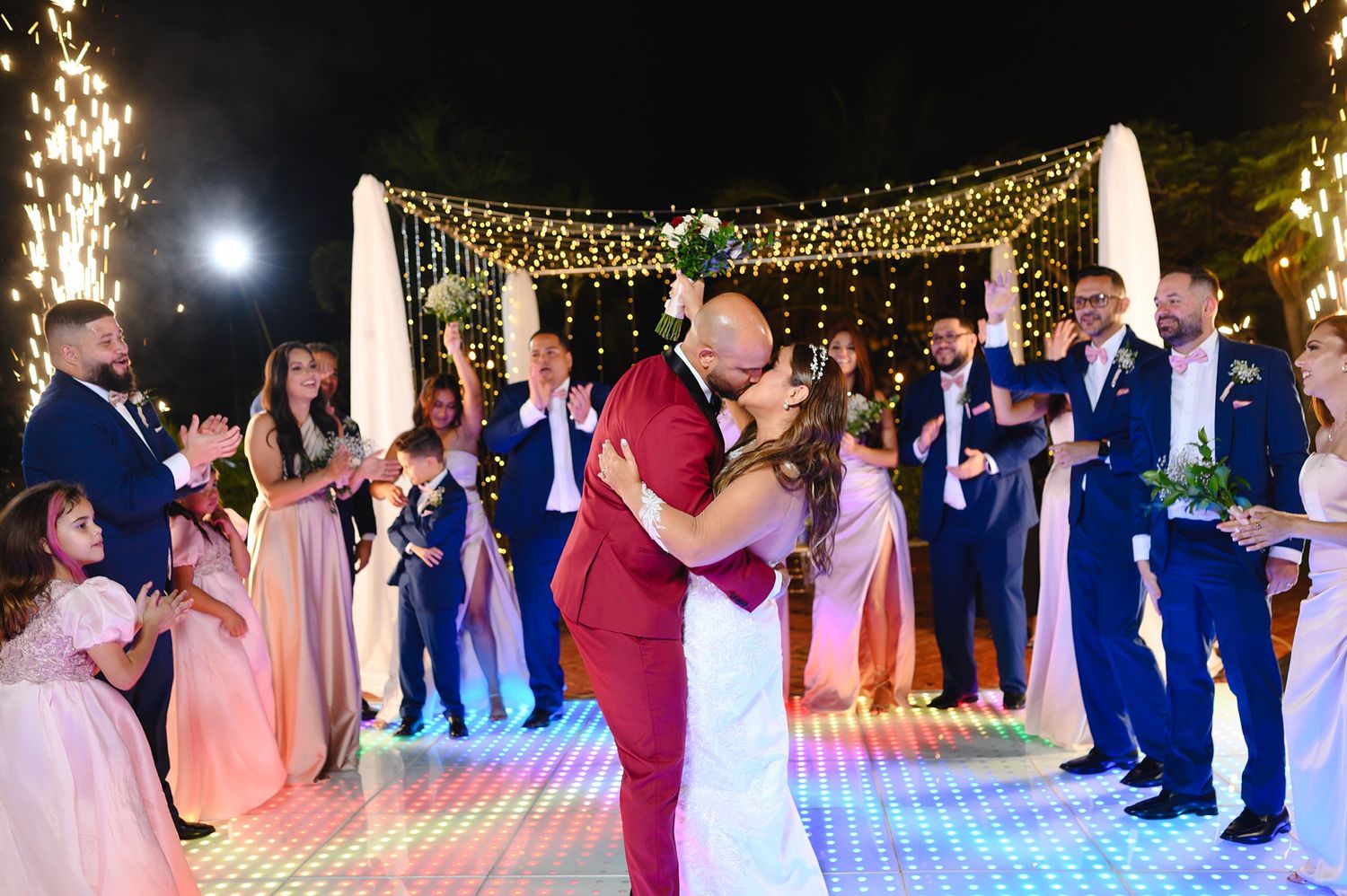 Bride and groom share first dance kiss at outdoor wedding reception with fairy lights and sparklers.