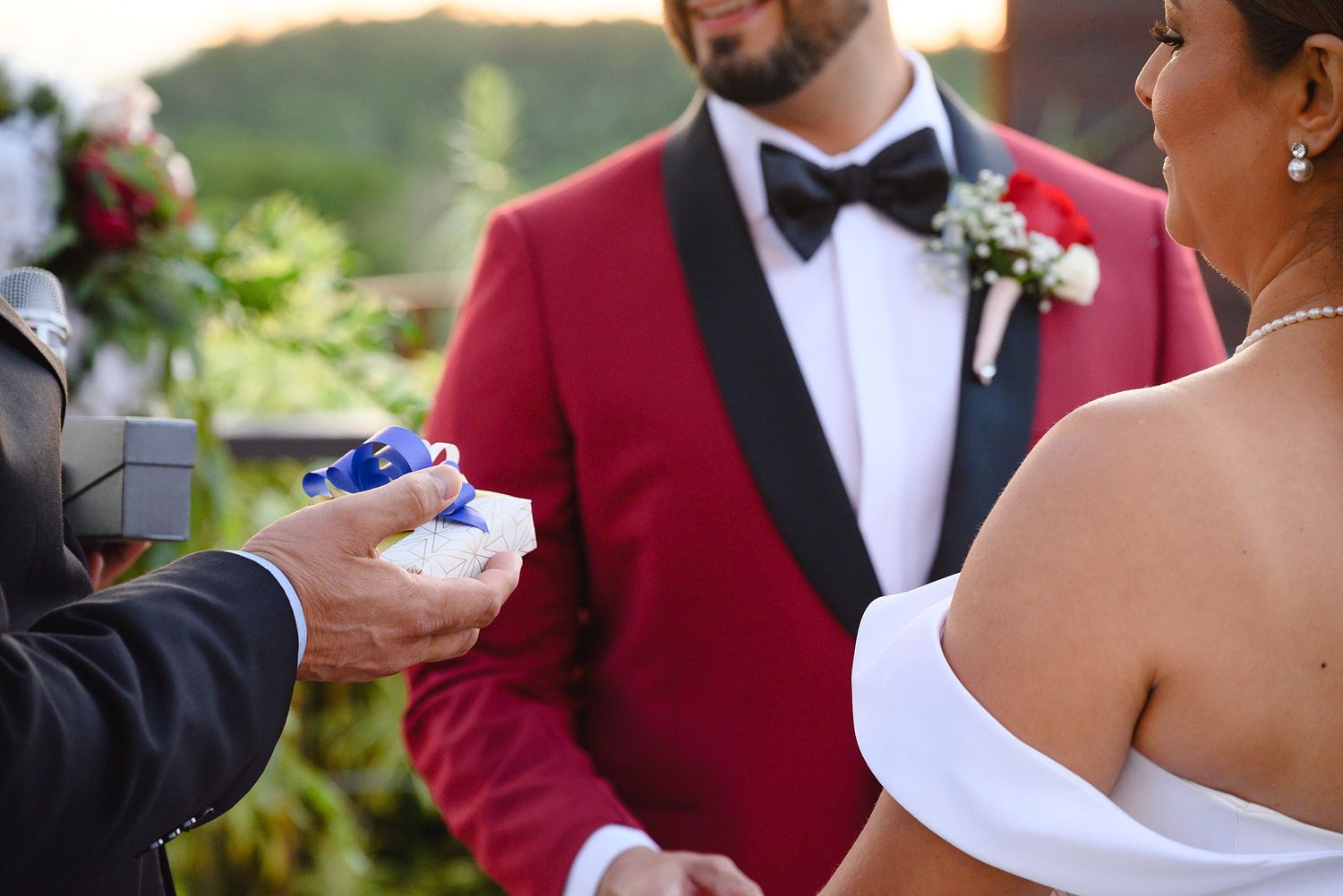 Groom in red tuxedo receives wedding rings during outdoor ceremony while bride stands nearby.