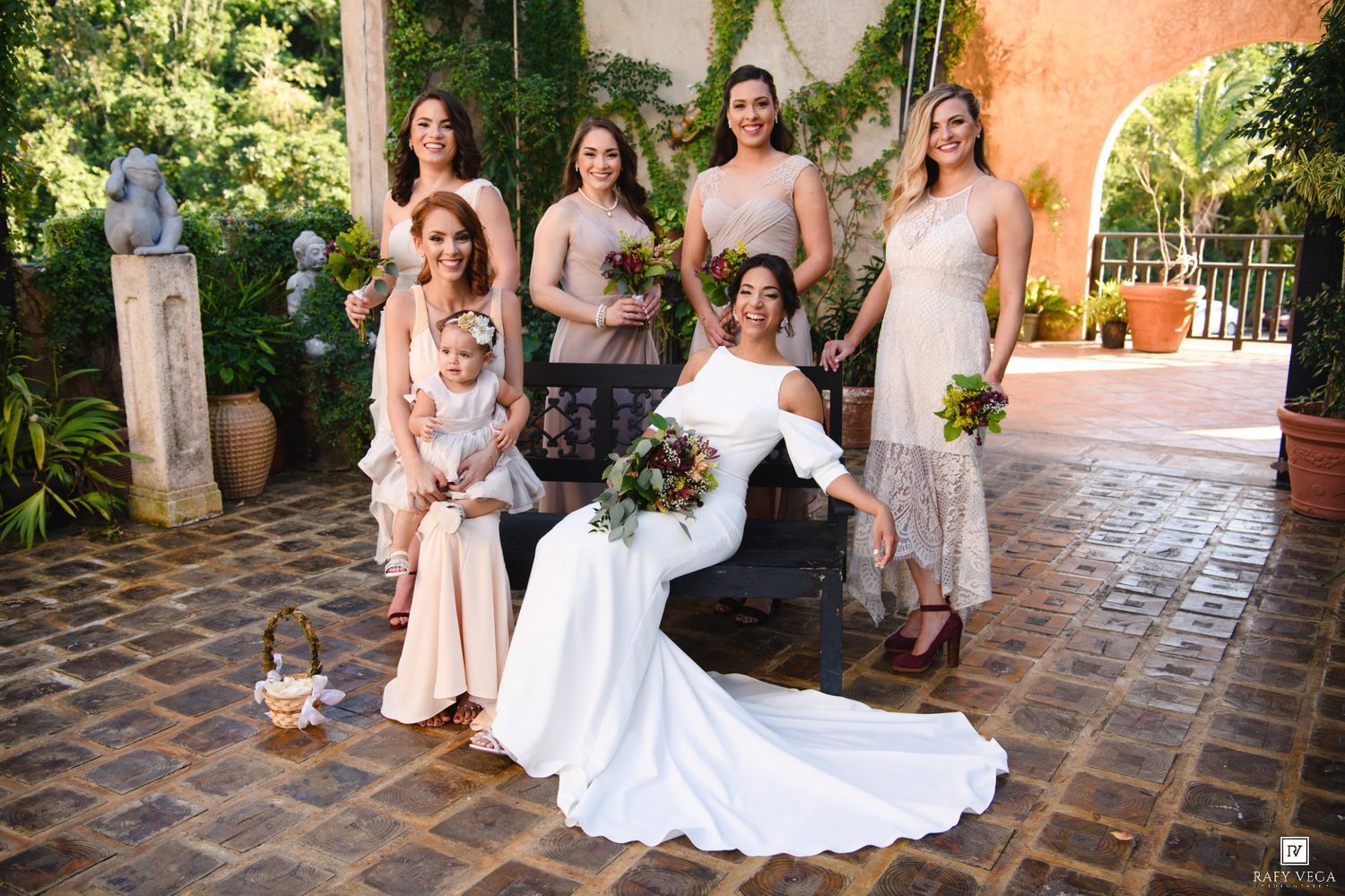 Bride in white gown poses with bridesmaids in blush dresses holding bouquet in elegant courtyard setting.
