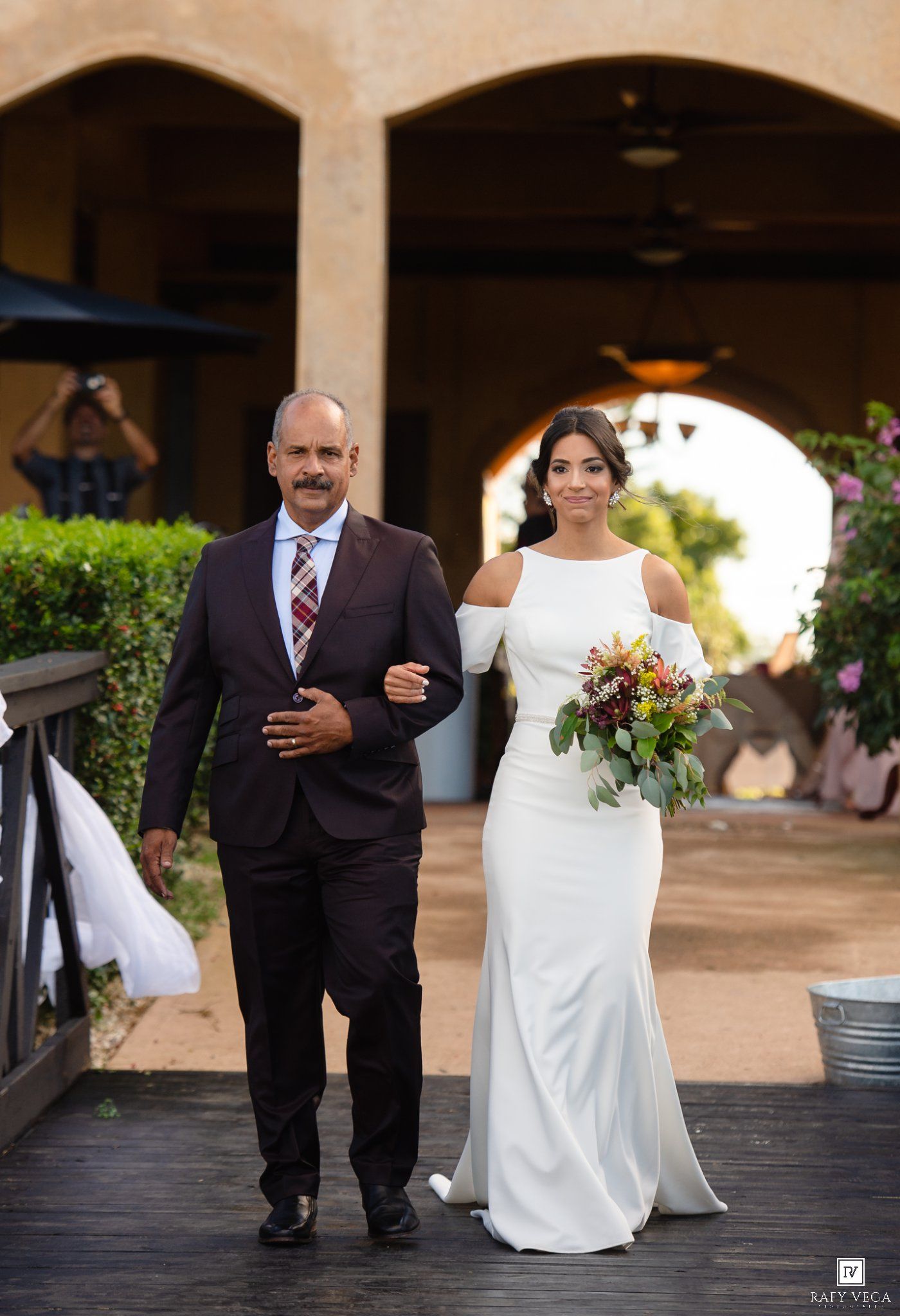 Bride in white off-shoulder gown walks down the aisle with her father in a dark suit at an outdoor wedding.