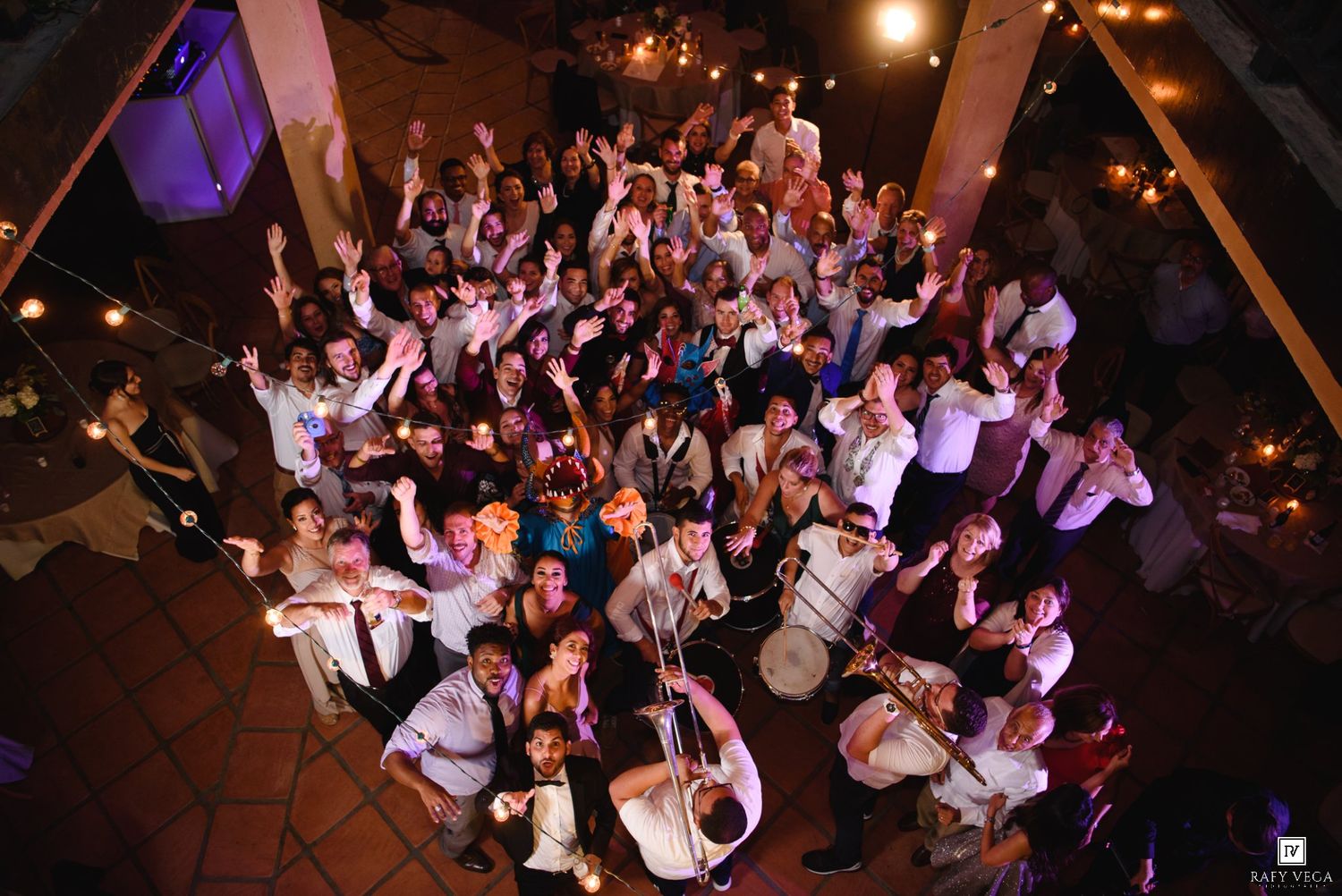 Aerial view of a large wedding reception group photo with live band performers under warm string lights.