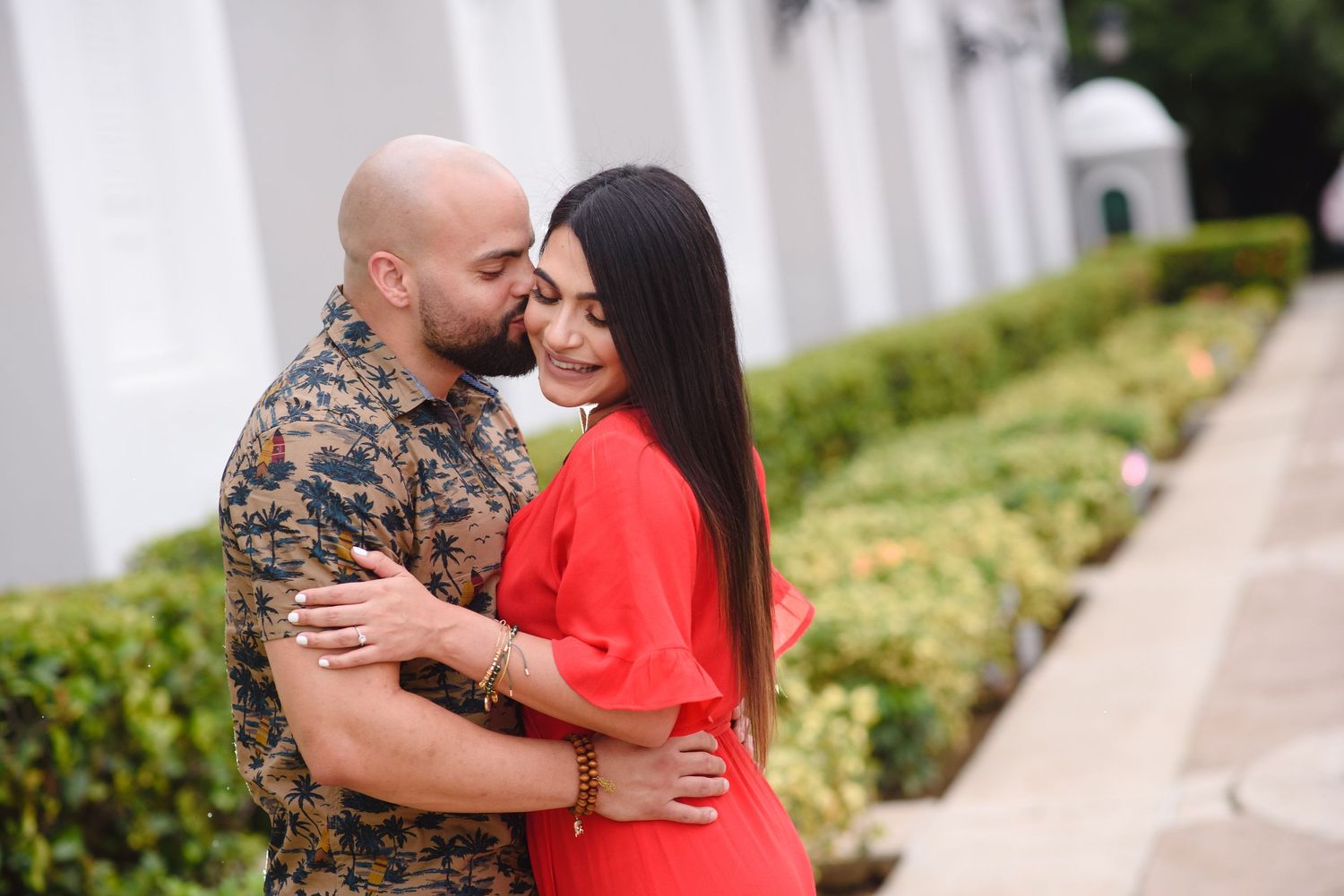 A bald man in floral shirt embraces a woman in red dress, sharing a romantic moment in a garden setting.