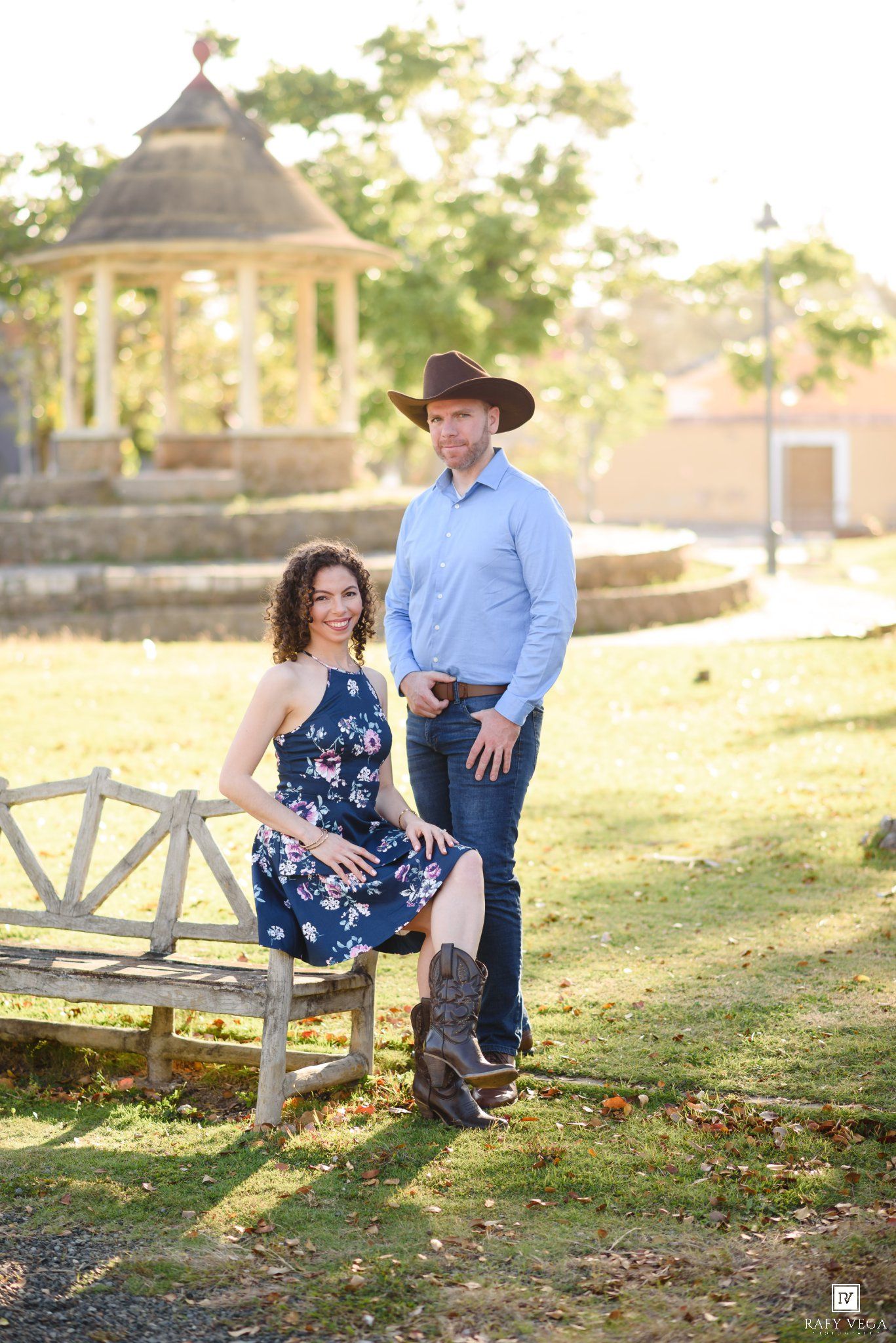 Couple posing outdoors near a rustic bench and gazebo, woman in floral dress, man in cowboy hat and jeans.