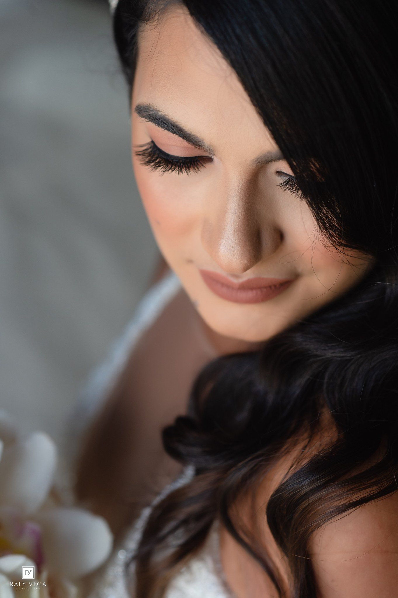 Close-up portrait of a bride with dark wavy hair, dramatic lashes, and soft glam makeup, looking down gracefully.