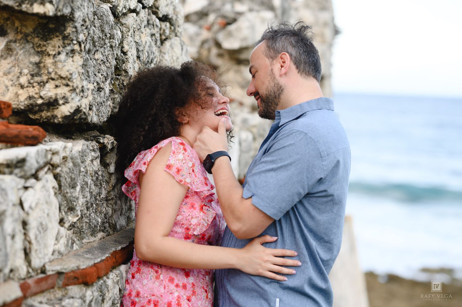 Couple laughing and embracing against a rustic stone wall by the ocean on a sunny day.