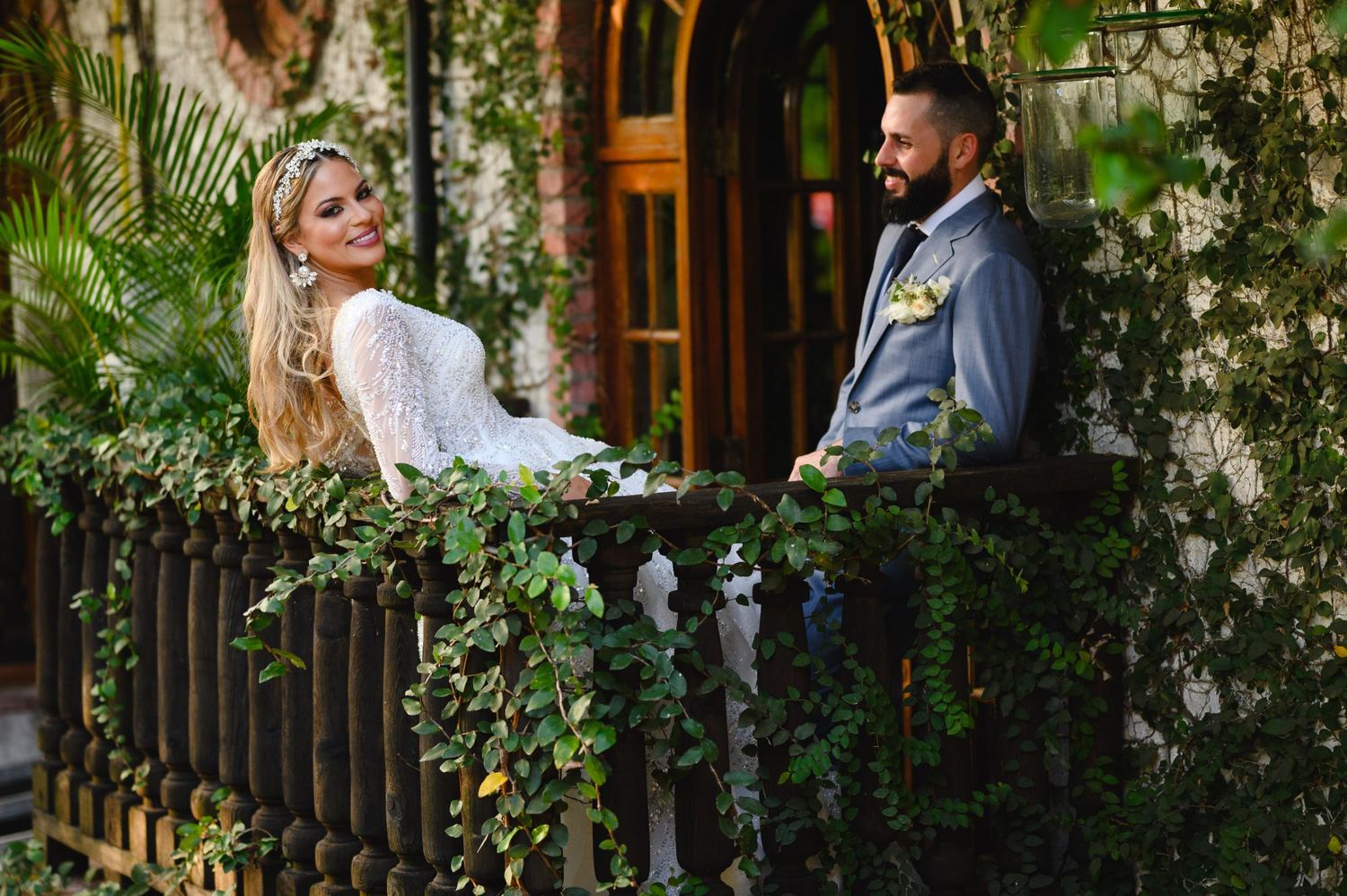 Bride and groom share a romantic moment on an ivy-draped balcony in a lush garden setting.