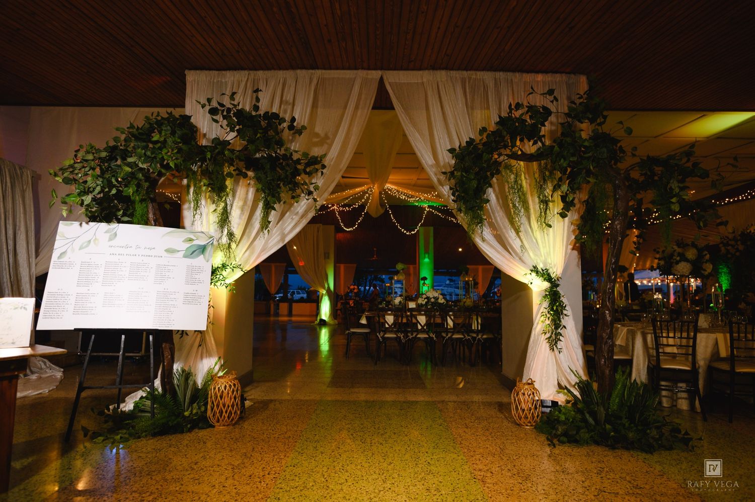 Elegant wedding reception entrance with white draped archway, lush greenery, warm lighting, and a seating chart display on the left.