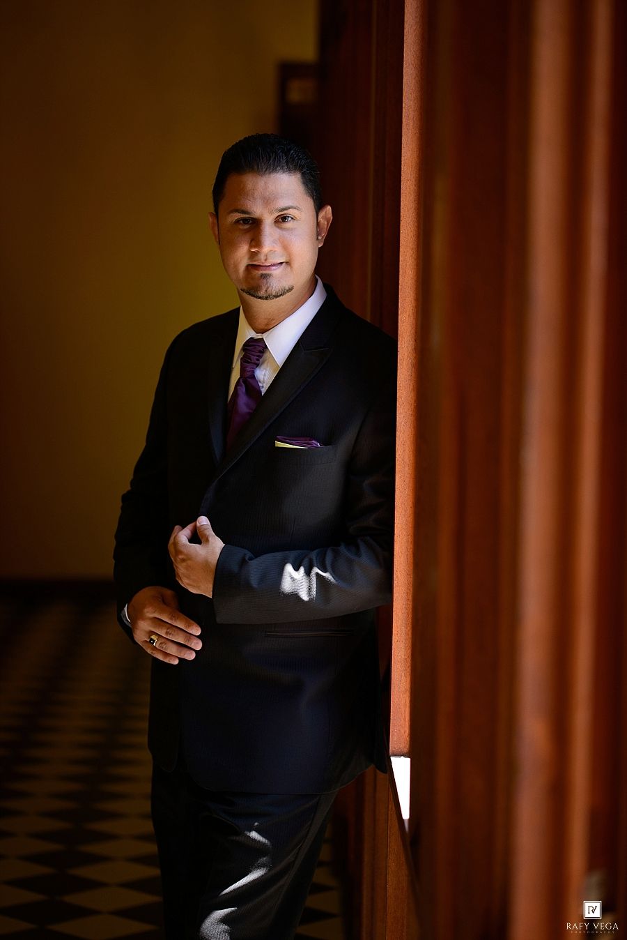 A well-dressed man in a black suit and purple tie poses confidently leaning against a wooden pillar in an elegant hallway.