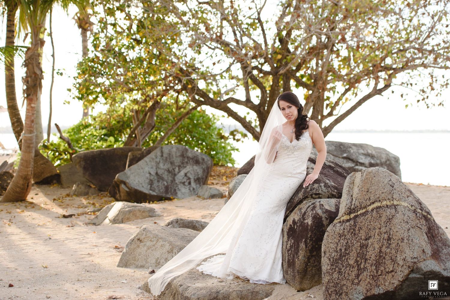 Elegant bride in fitted white wedding dress sitting on rocks at tropical beach with palm trees.