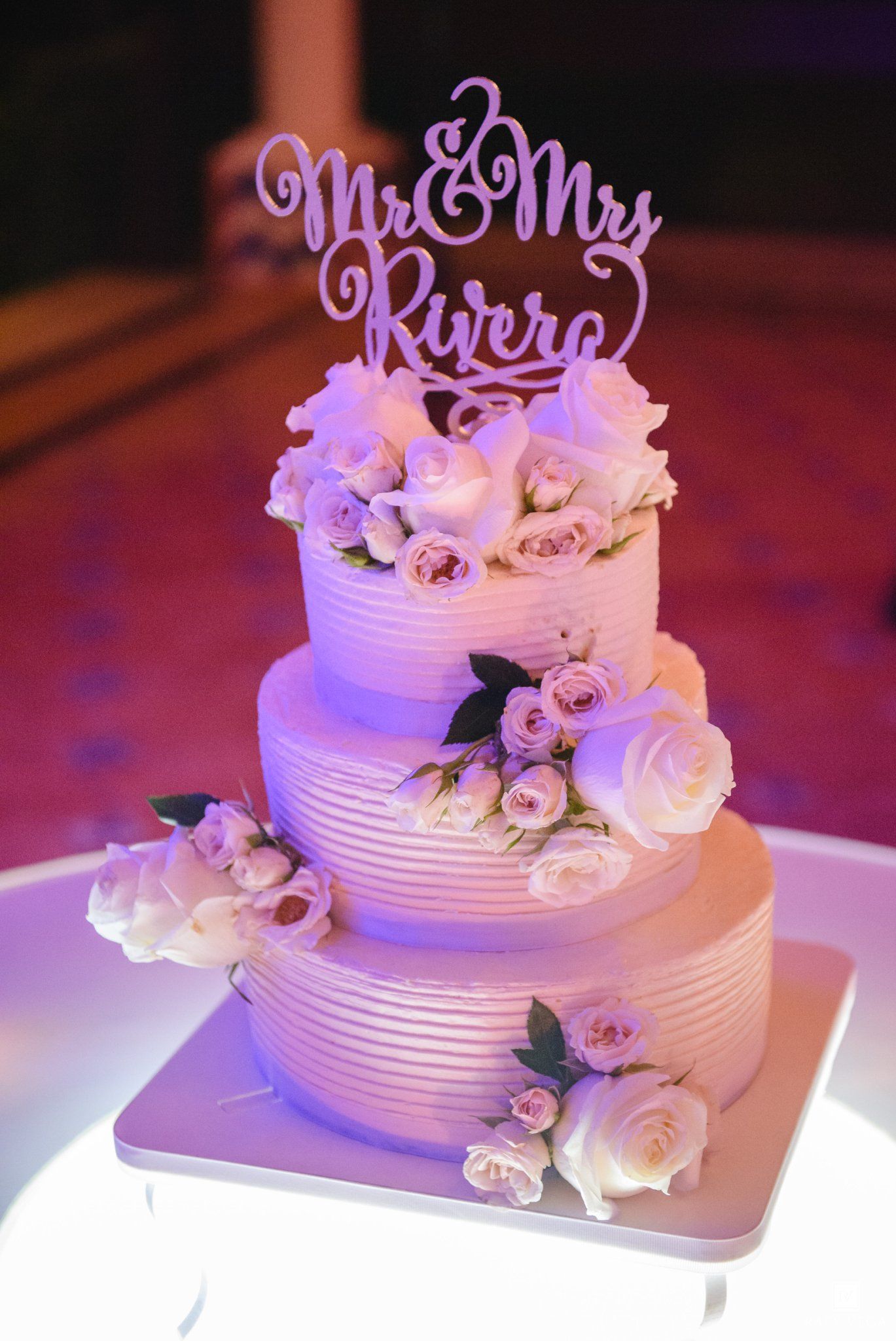Three-tier white wedding cake with pink roses and Mr & Mrs Rivera cake topper under purple lighting.