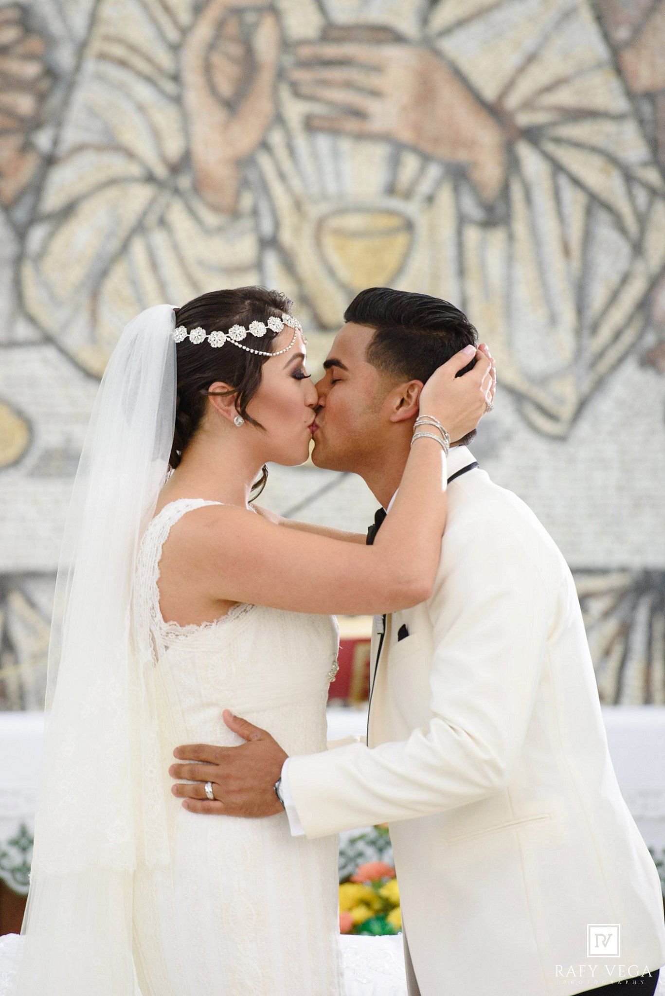 Bride and groom share first kiss at church altar, bride in white gown with veil, groom in white tuxedo.