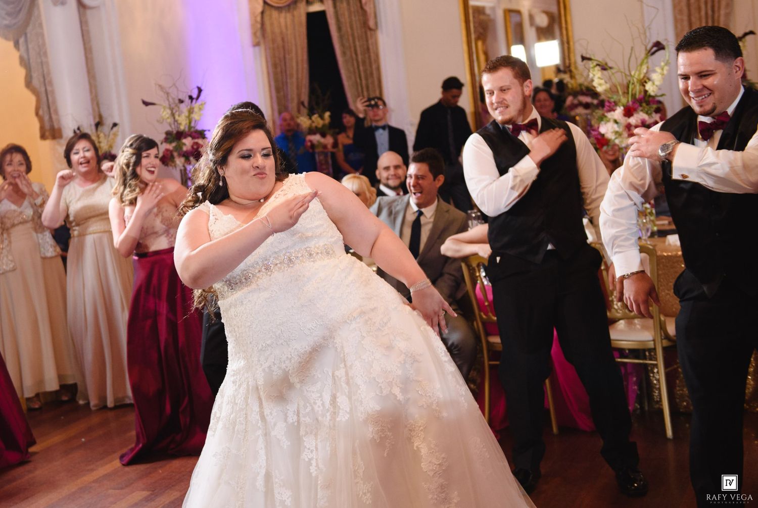 Bride in white gown dancing energetically with groom in tuxedo at elegant wedding reception ballroom.