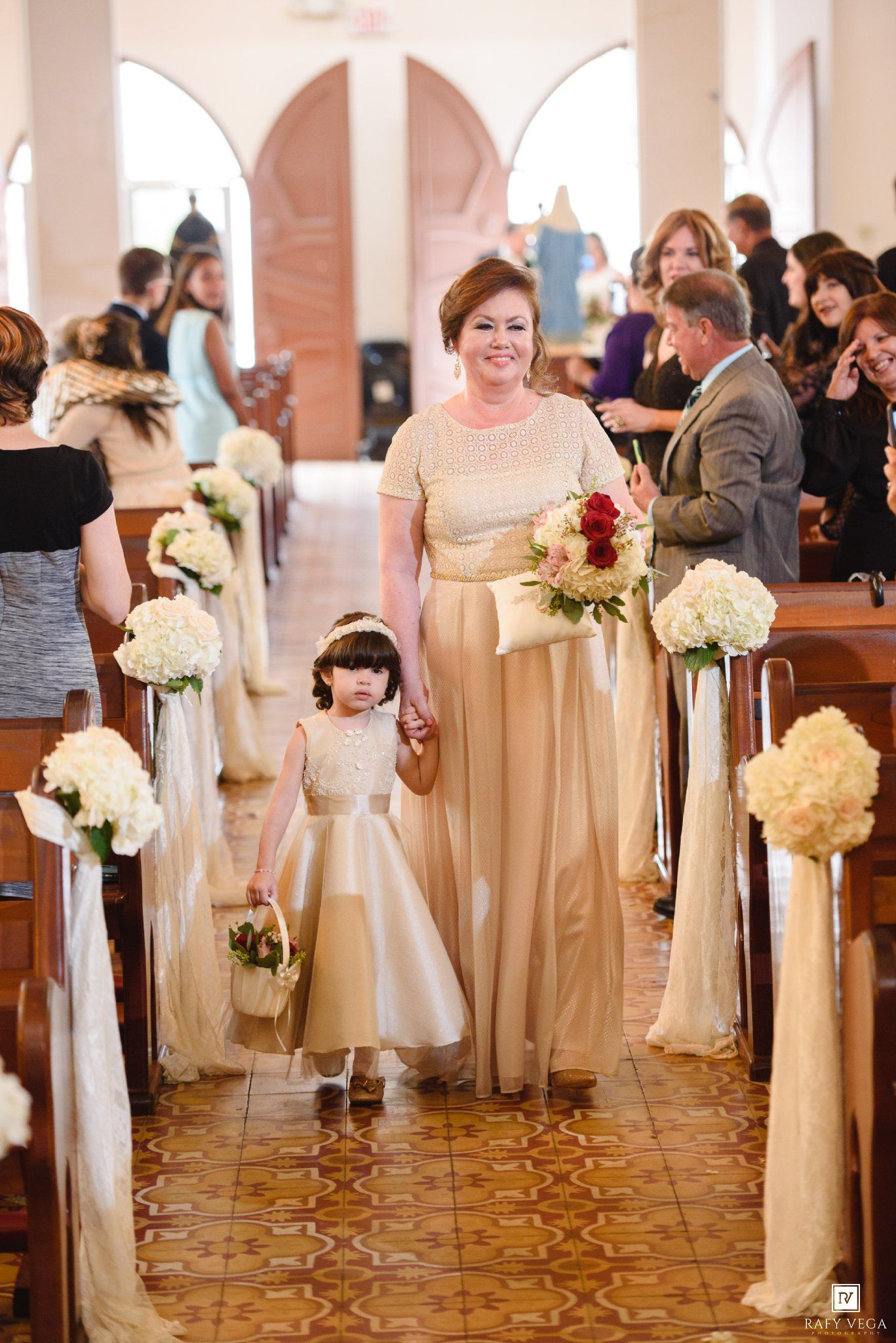 Elegant woman in gold gown walks flower girl down church aisle decorated with white floral arrangements.