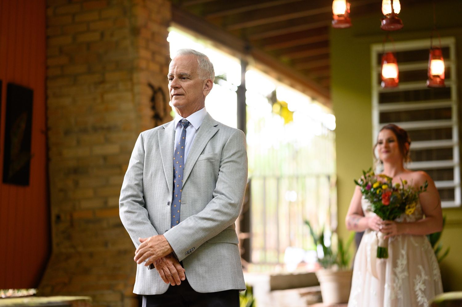 Older man in gray suit stands at outdoor wedding ceremony as bride in white dress holds yellow bouquet in background.