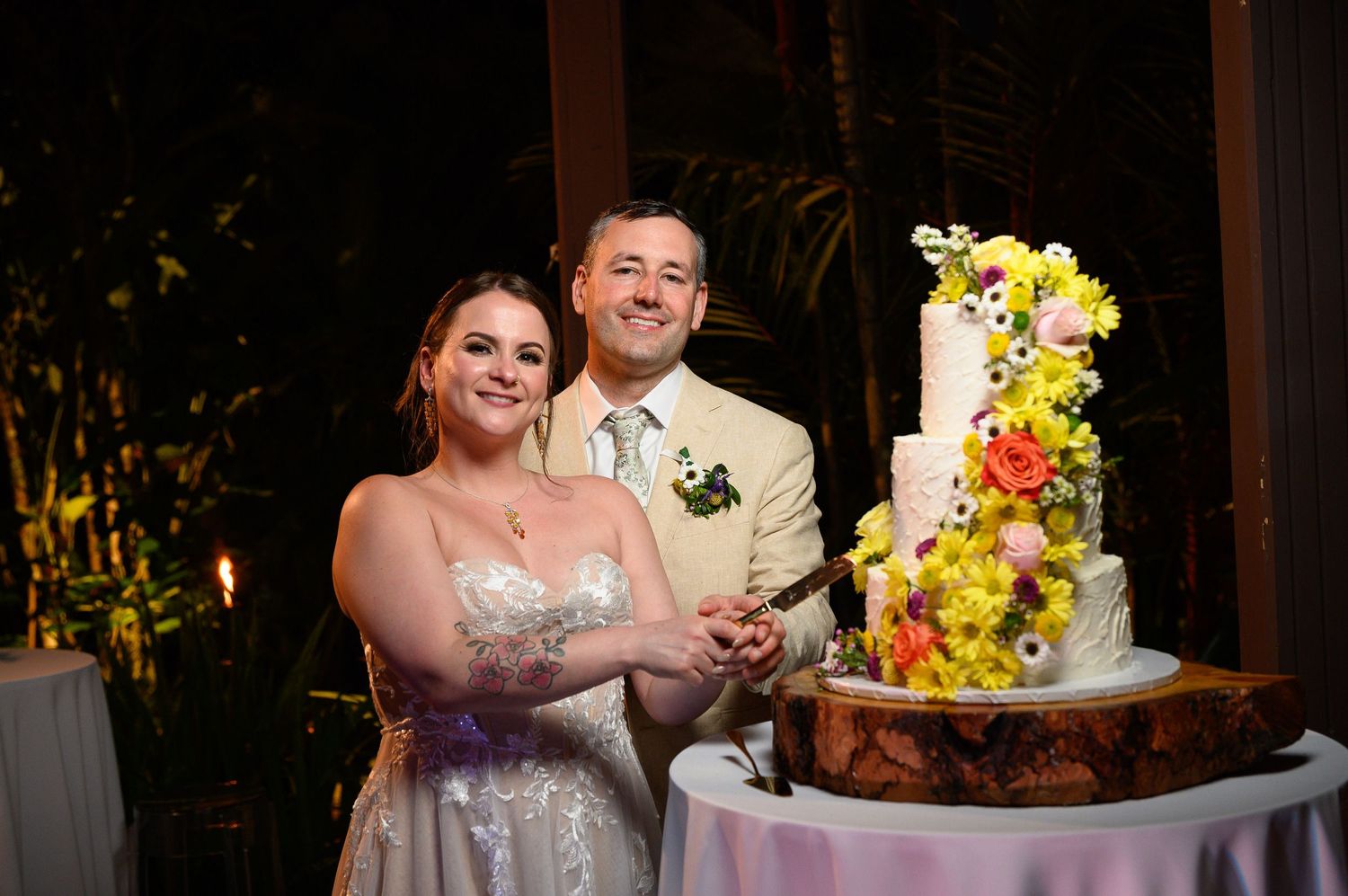 Bride and groom smiling while cutting their floral wedding cake decorated with yellow and orange flowers.