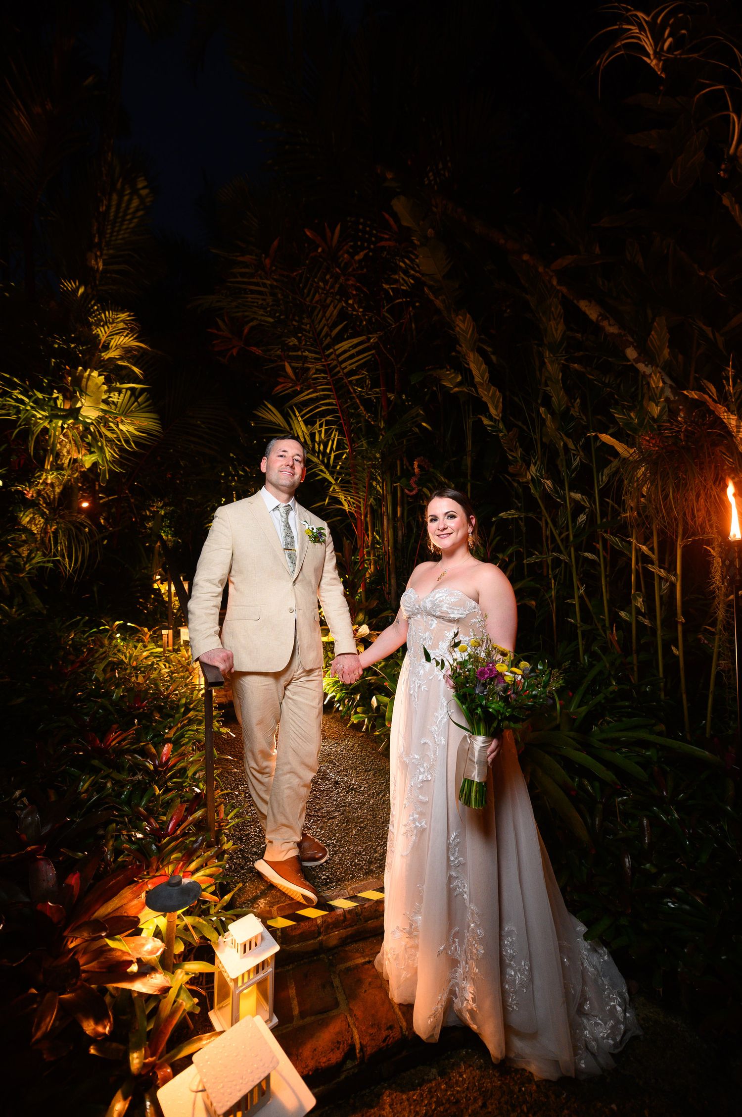 Bride and groom hold hands in a lush tropical garden at night, illuminated by warm torchlight.
