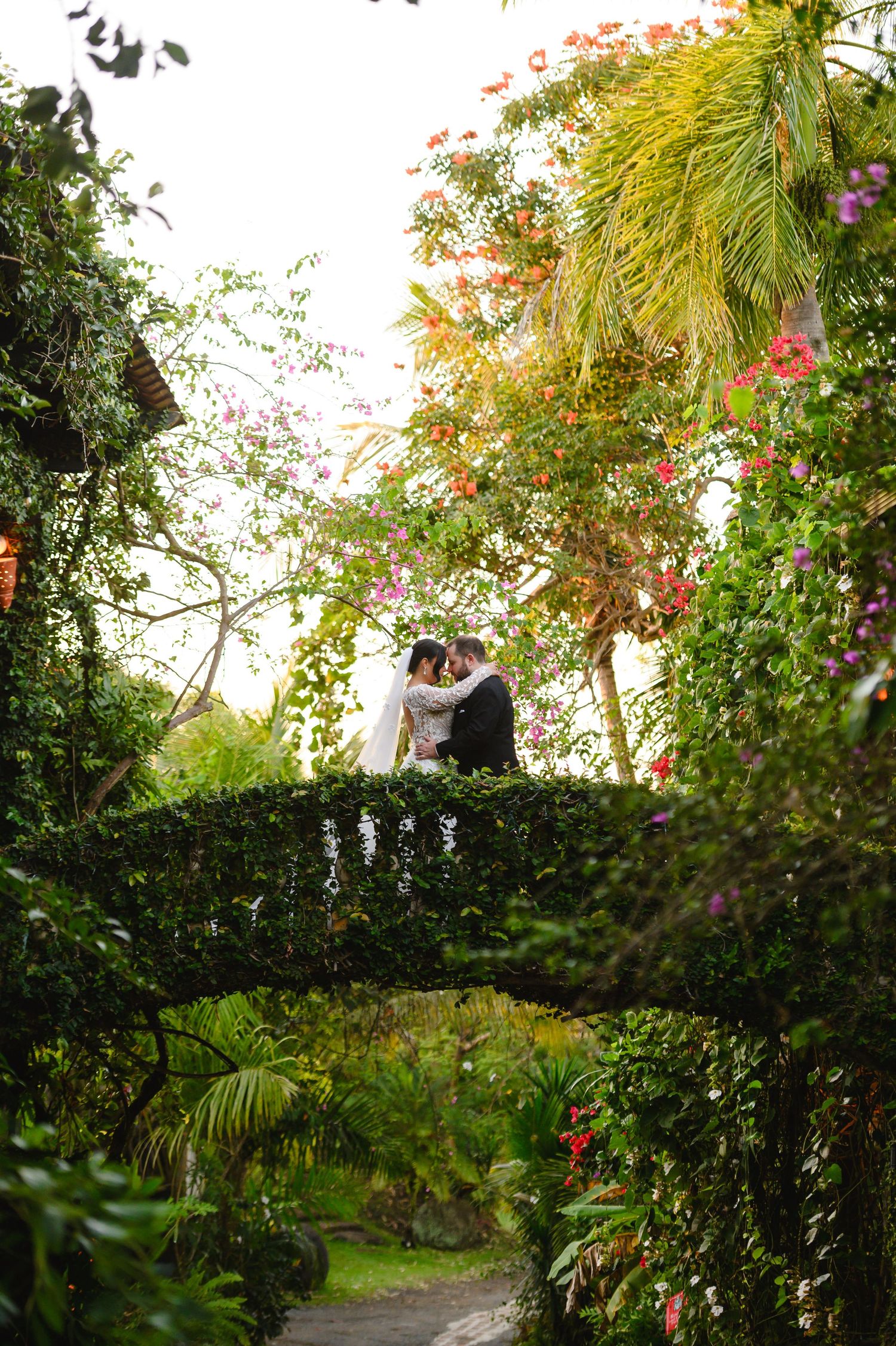 Bride and groom share a romantic moment on an ivy-covered stone bridge surrounded by lush tropical gardens and colorful flowers.