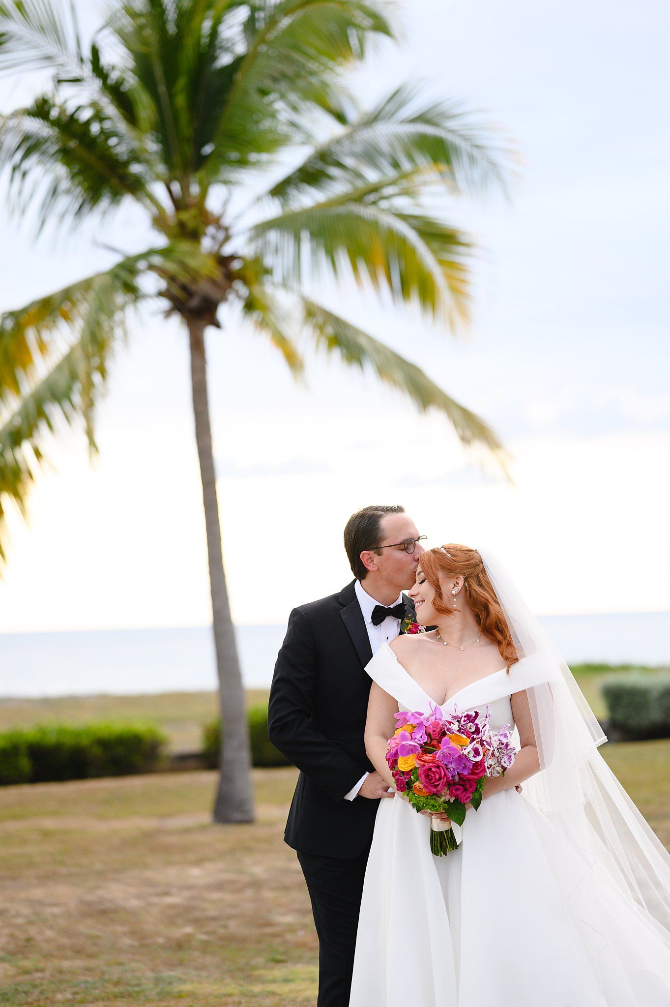 Bride and groom share a romantic kiss near a tropical palm tree with ocean views, bride holding vibrant pink bouquet.