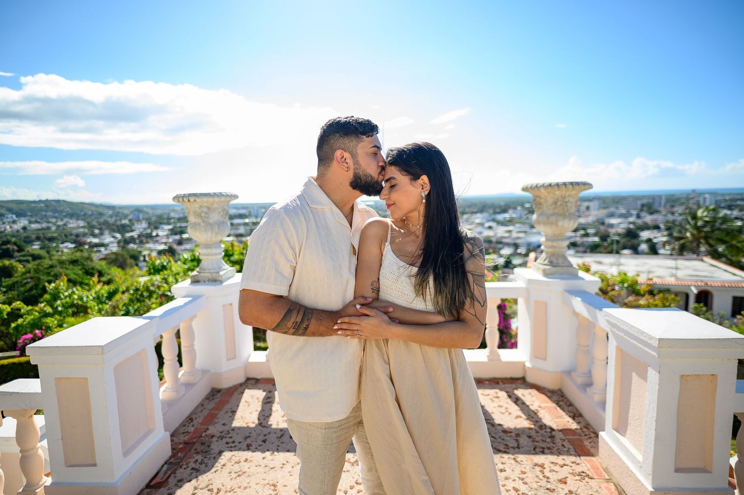Couple embracing and kissing on a white balcony terrace with a scenic city skyline view on a sunny day.