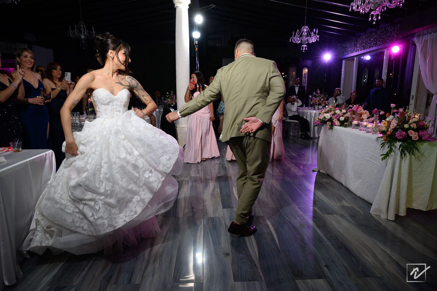 Bride in a white lace gown and groom in an olive suit walk across a dance floor at an elegant wedding reception with floral centerpieces.