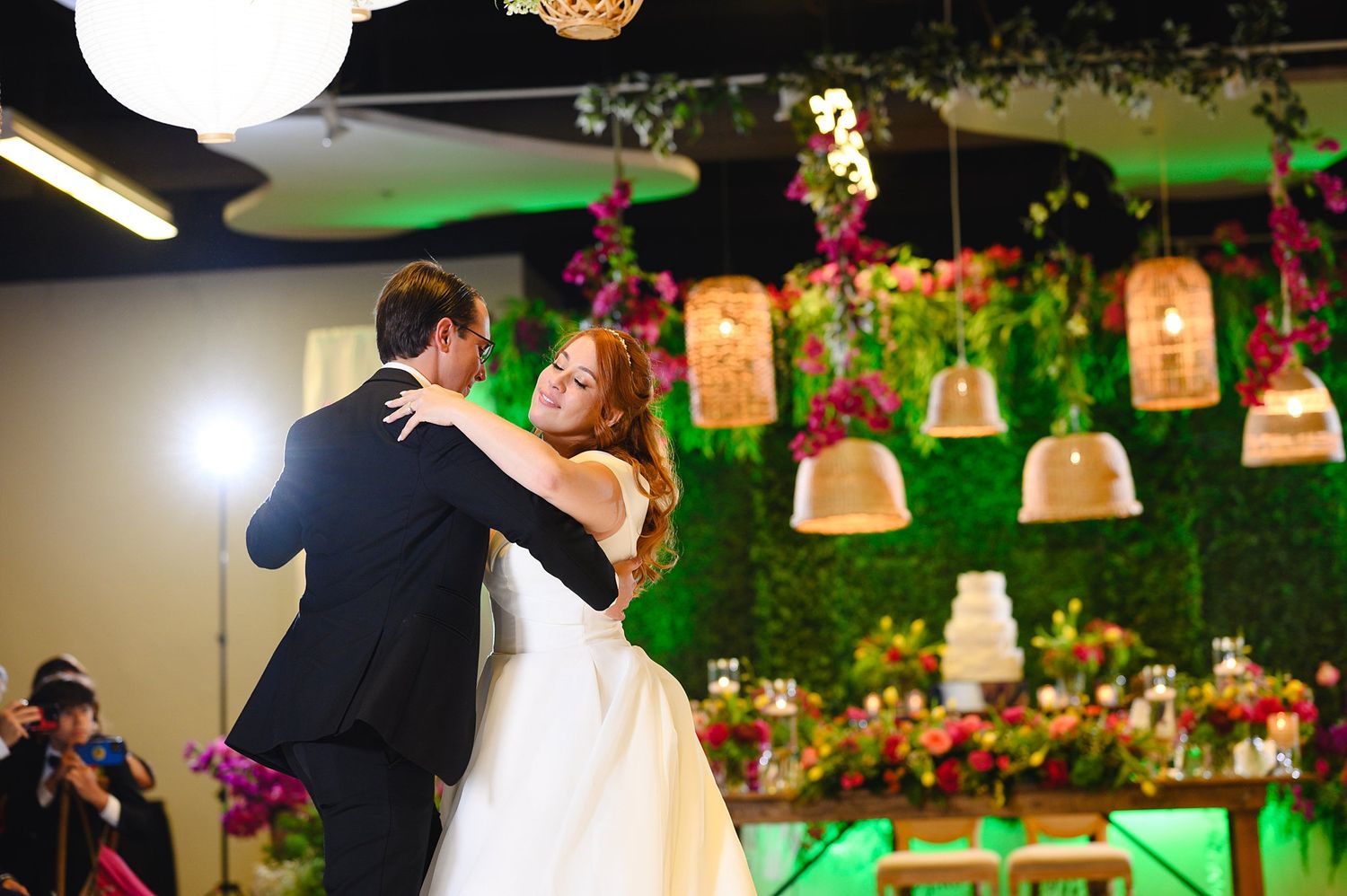 Bride and groom share their first dance at a wedding reception with floral backdrop and elegant cake display.