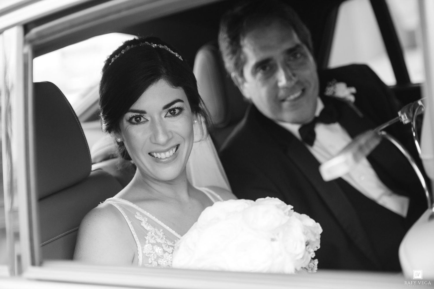 Black and white photo of a smiling bride in a wedding dress sitting in a car with a man in a suit beside her.