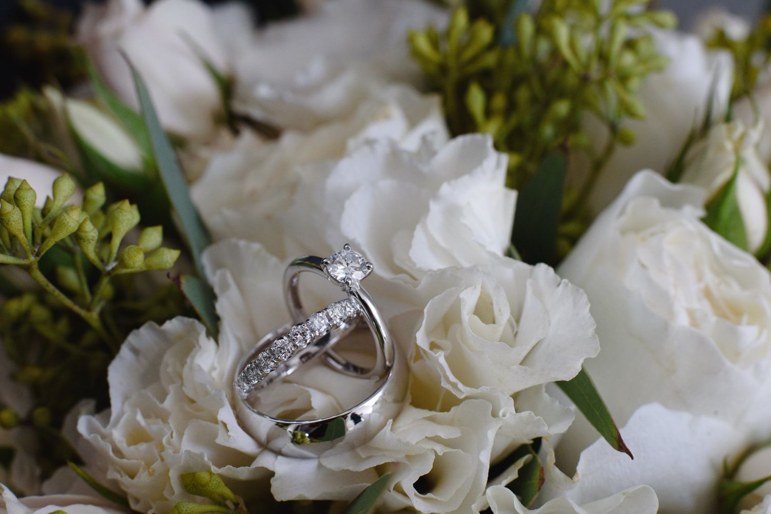 Two silver wedding rings with diamonds rest on white roses and green floral bouquet.