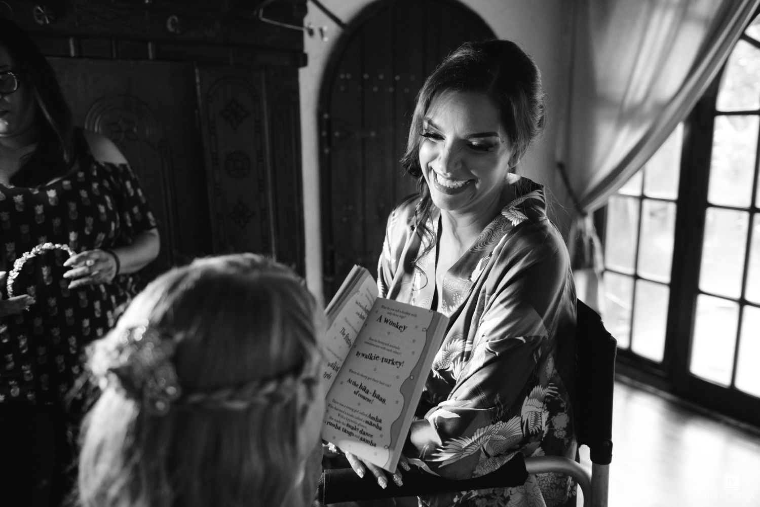 Black and white photo of a smiling woman showing a book to a bride with a braided updo at a wedding.