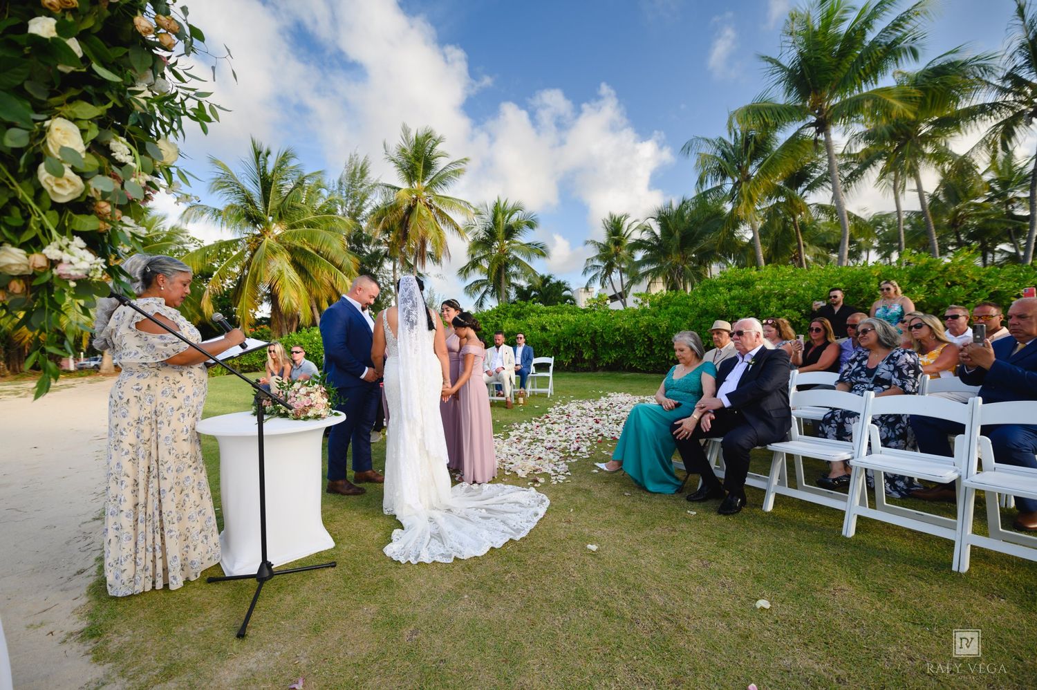Outdoor beach wedding ceremony with couple at altar, officiant, and guests seated on white chairs surrounded by palm trees.