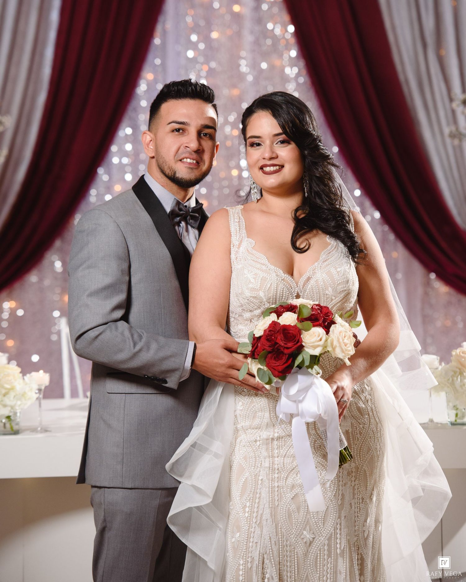 A bride in a beaded gown holds red roses beside a groom in a gray tuxedo before a sparkling backdrop.