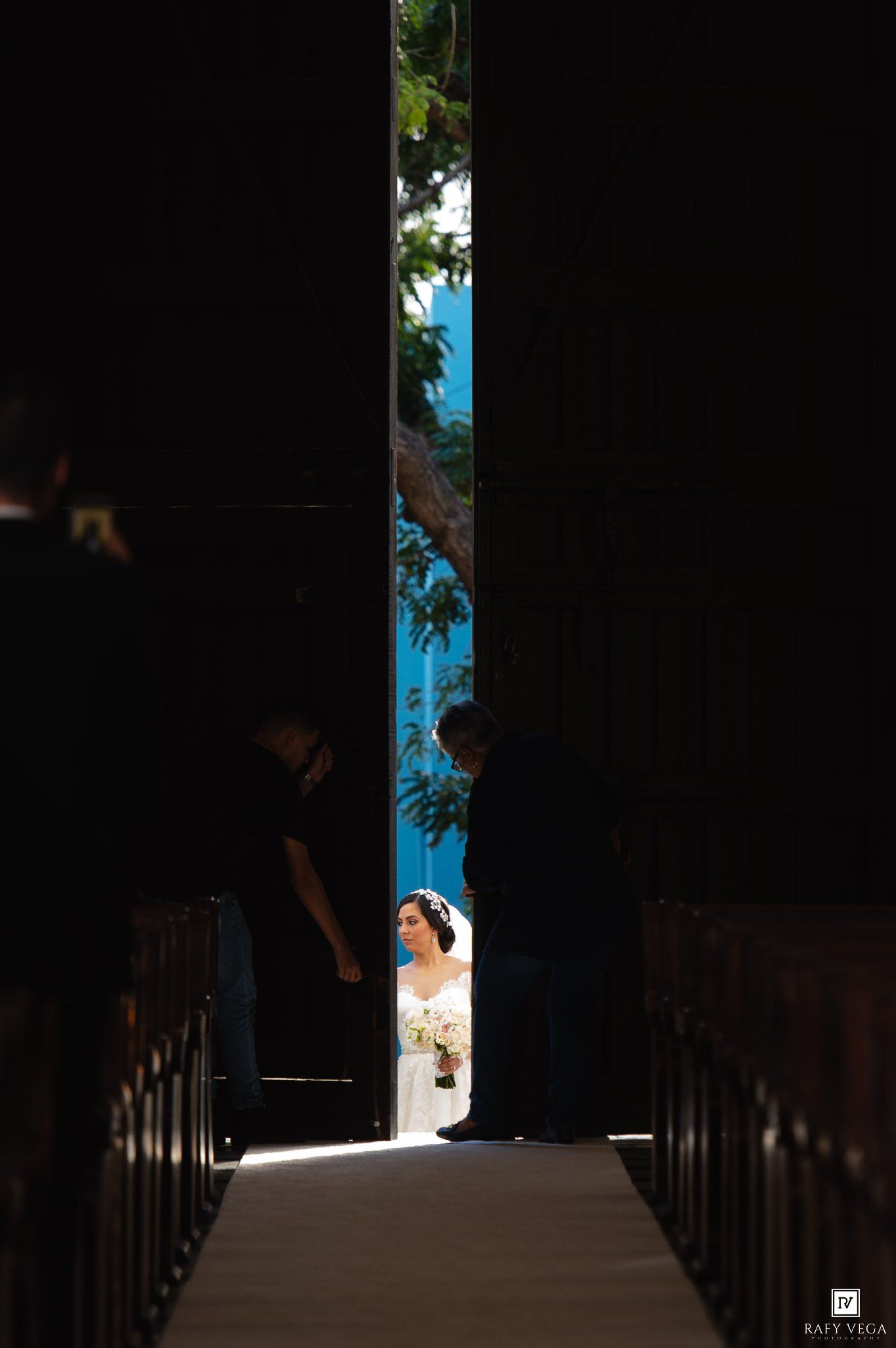 Bride waiting at church entrance seen through narrow doorway gap before wedding ceremony.