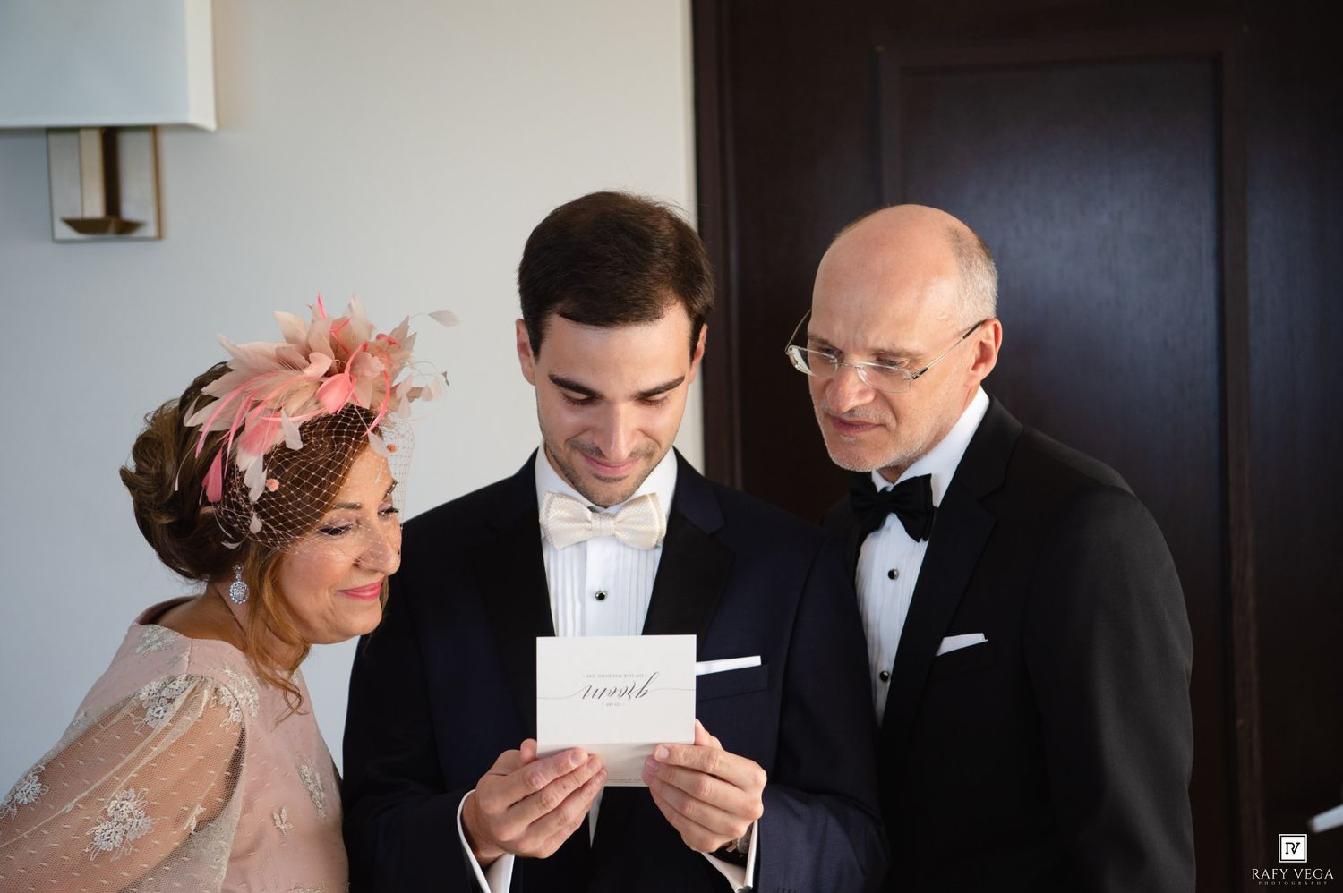 Groom in tuxedo reads a card with elegantly dressed parents beside him before wedding ceremony.