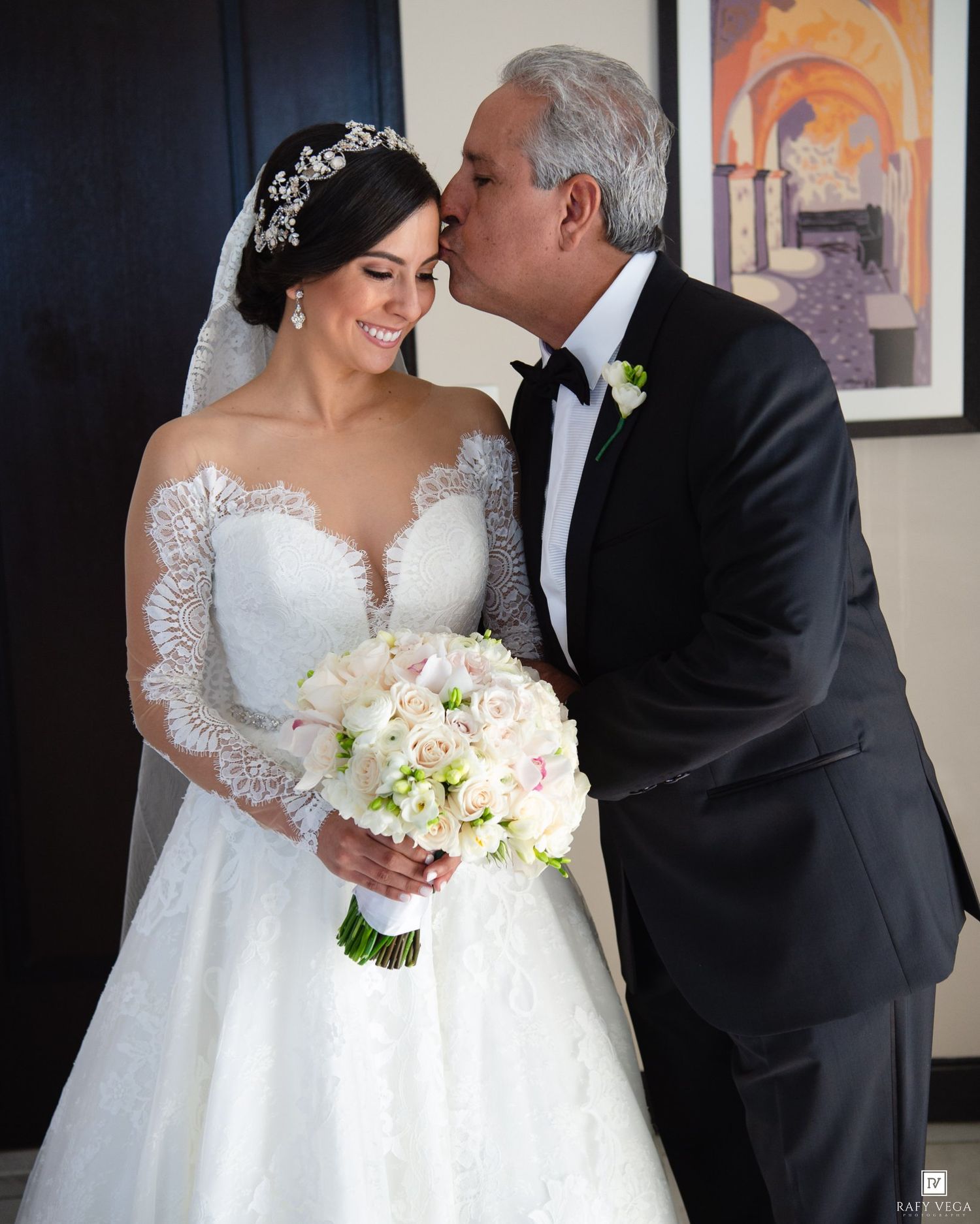 A bride in a lace gown holds a bouquet as an older man in a tuxedo kisses her cheek on her wedding day.