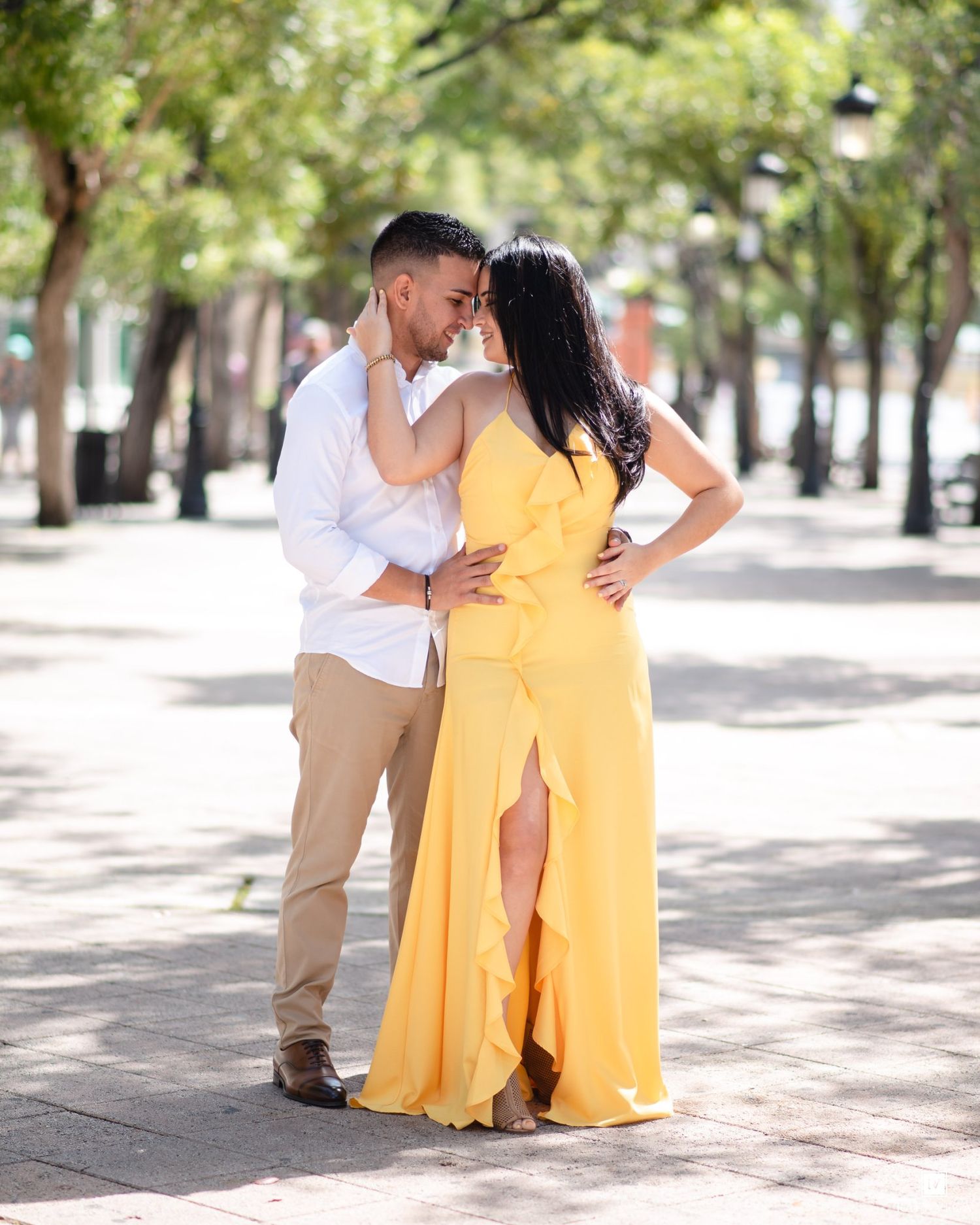 Couple in romantic pose outdoors, woman in yellow maxi dress, man in white shirt and khakis, tree-lined walkway.