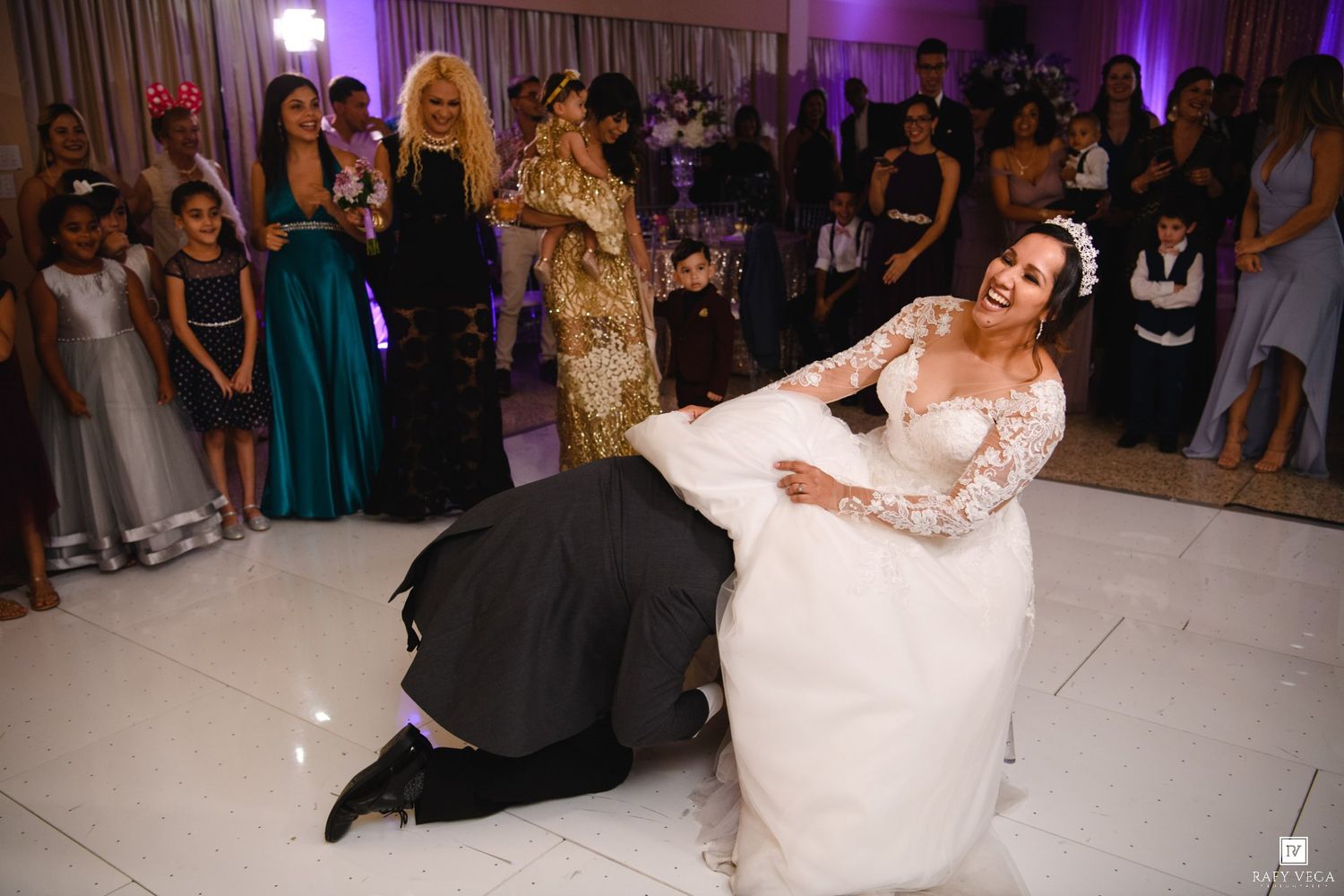 Bride laughing on dance floor as groom kneels before her during lively wedding reception celebration.